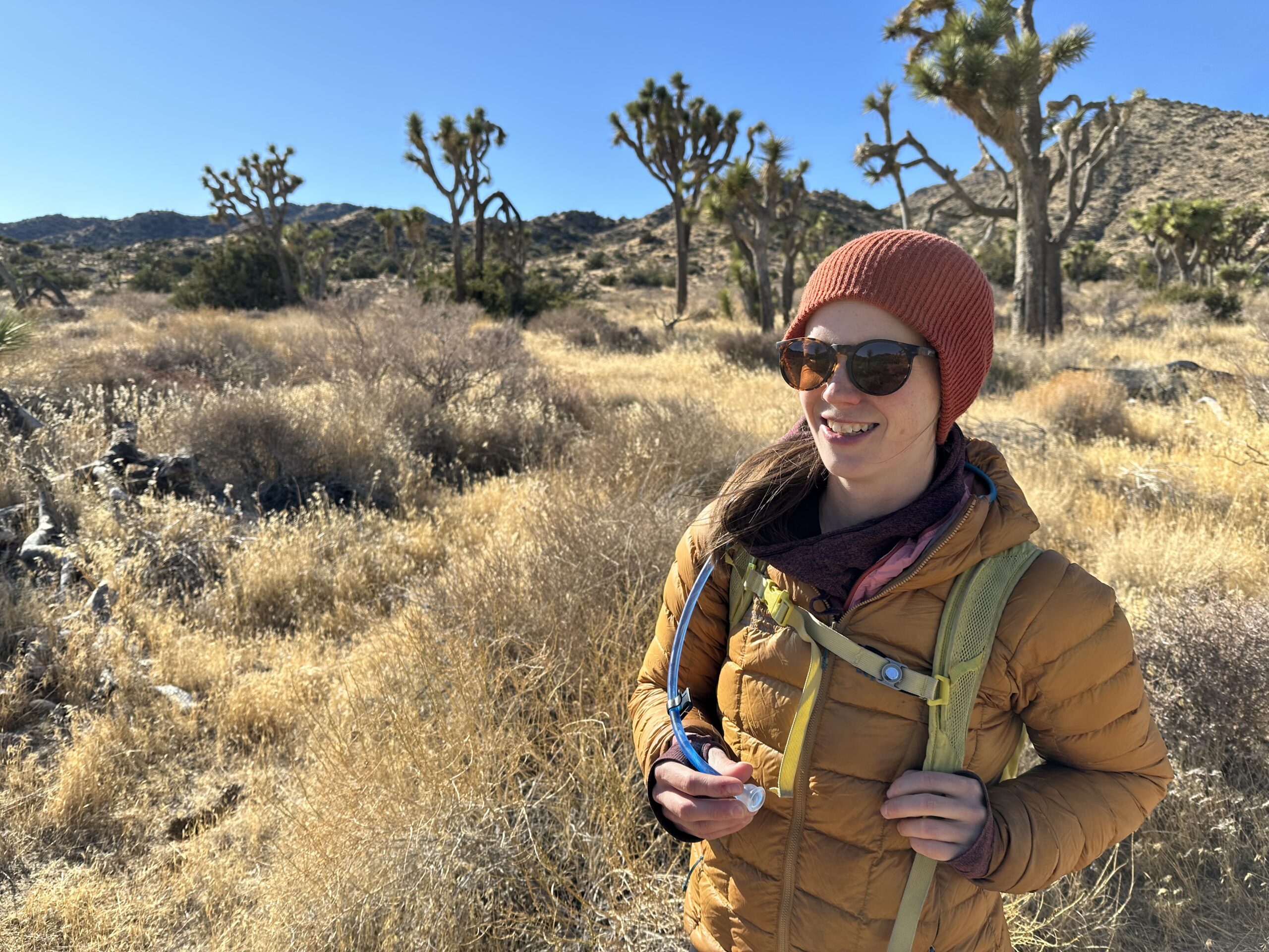 A hiker smiles on a trail with Joshua Trees in the background, holding the bite valve of the Platypus Hoser Reservoir.