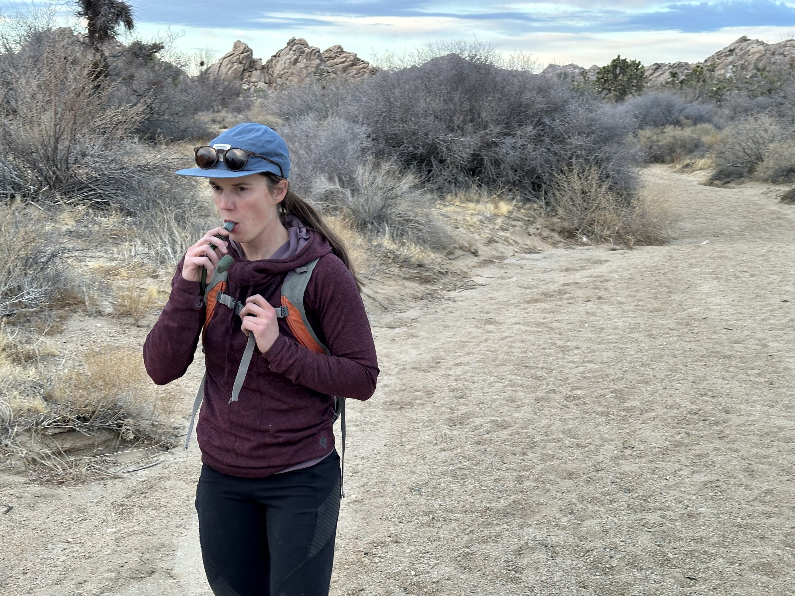 A hiker drinks from the bite valve of the Cherainti Bladder while hiking in a sandy wash in Joshua Tree National Park. Joshua Trees and granite rock formations are in the background.