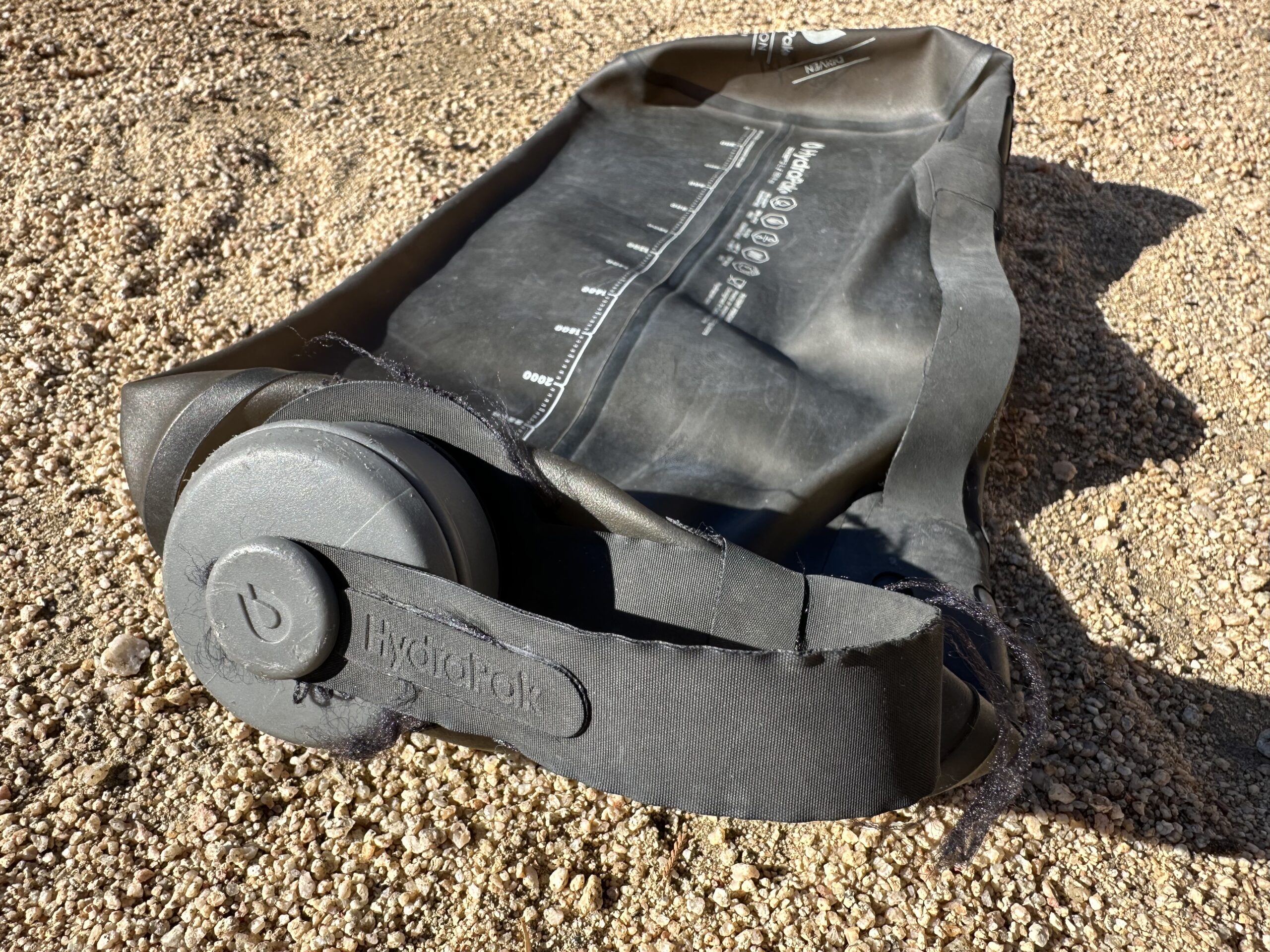 A close up photo shows an empty HydraPak Seeker water bag laying on the sand. The fabric strap connecting the twist cap to the bladder is starting to fray.
