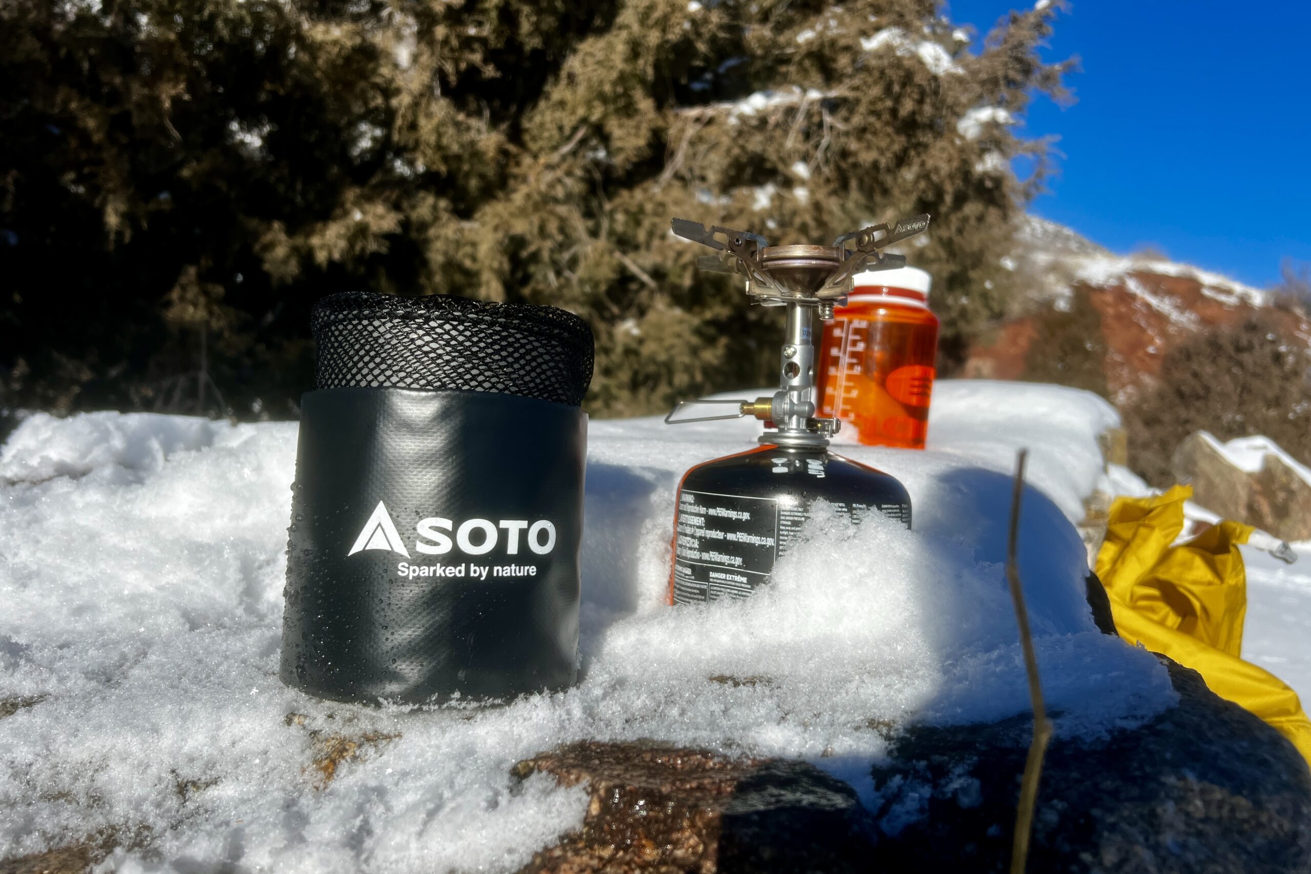The Soto thermostack combo sits packed in its stuff sack on snow next to a stove and nalgene bottle with red rocks in the background.