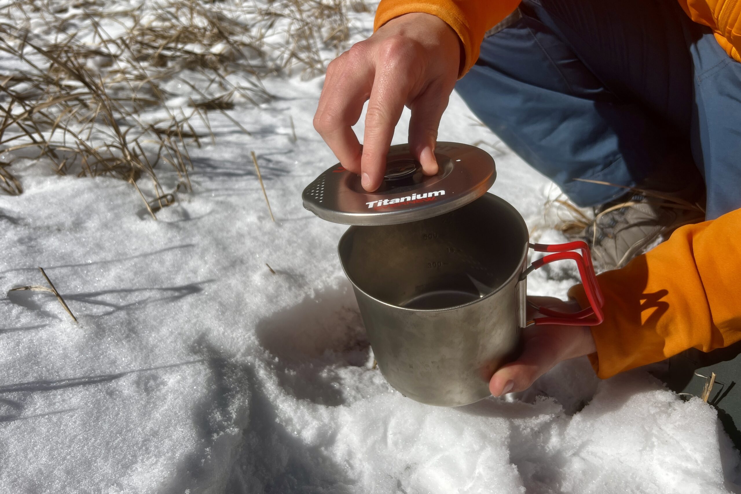 A hiker removes the lid of the evernew pasta pot against a snowy background.