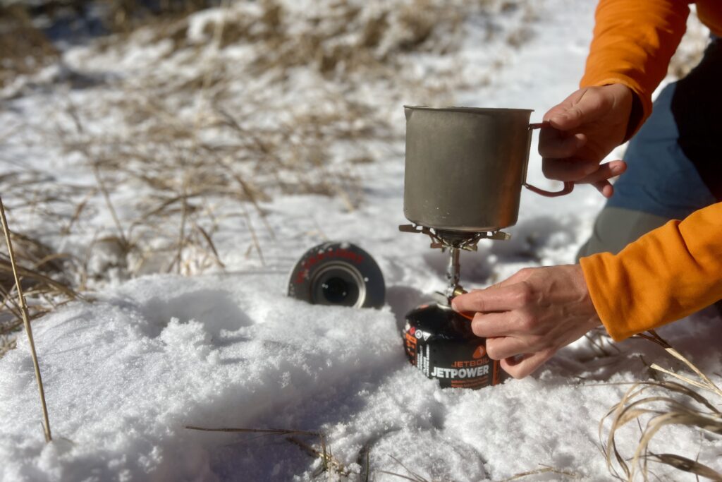 A close up of a hiker holding the evernew pasta pot on a stove with one hand and turning the stove on with the other. The lid of the pot is sticking out of snow.