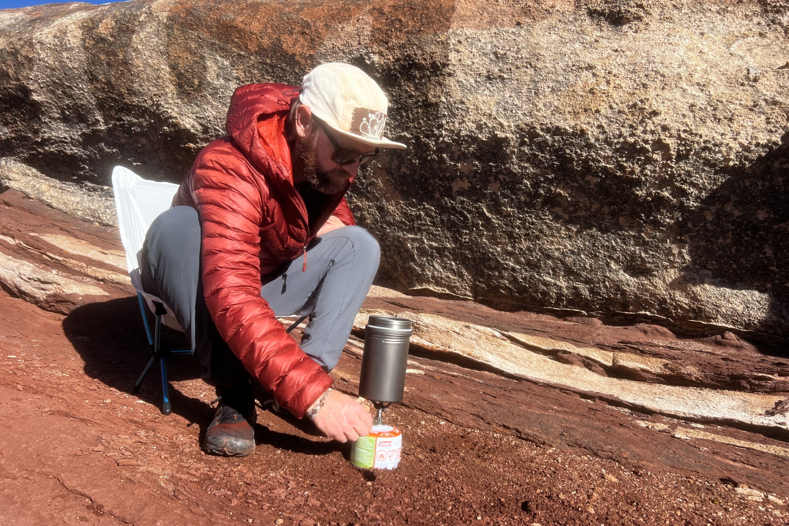 A hiker sits in a chair and lights a backpacking stove with a pot on top against a red rocks background.