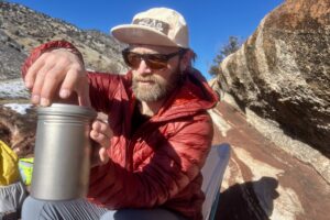 A hiker screws off the led to a titanium backpacking pot with rocks in the background.
