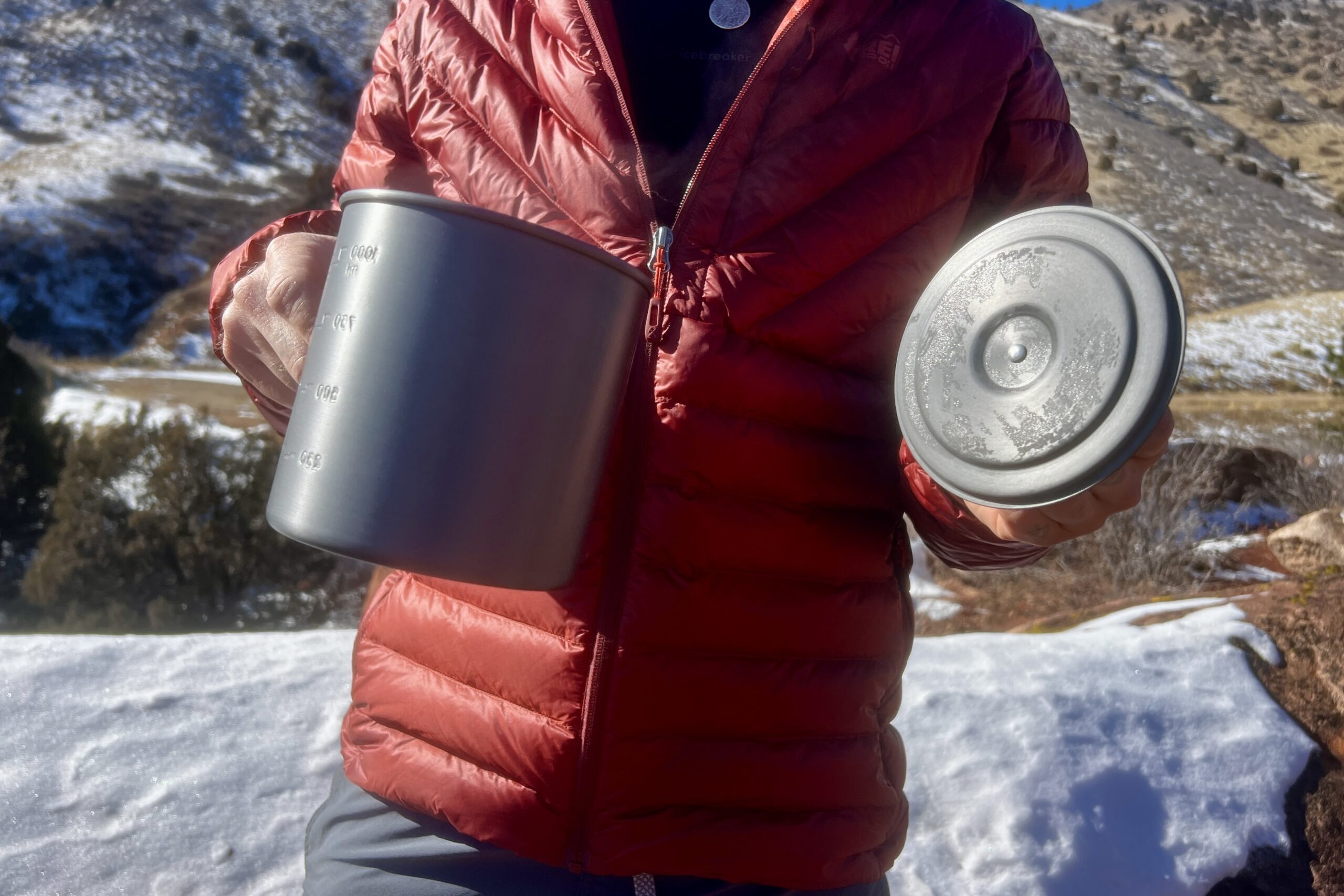 A hiker holds the halulite boiler in one hand and the lid in the other with a snowy background.