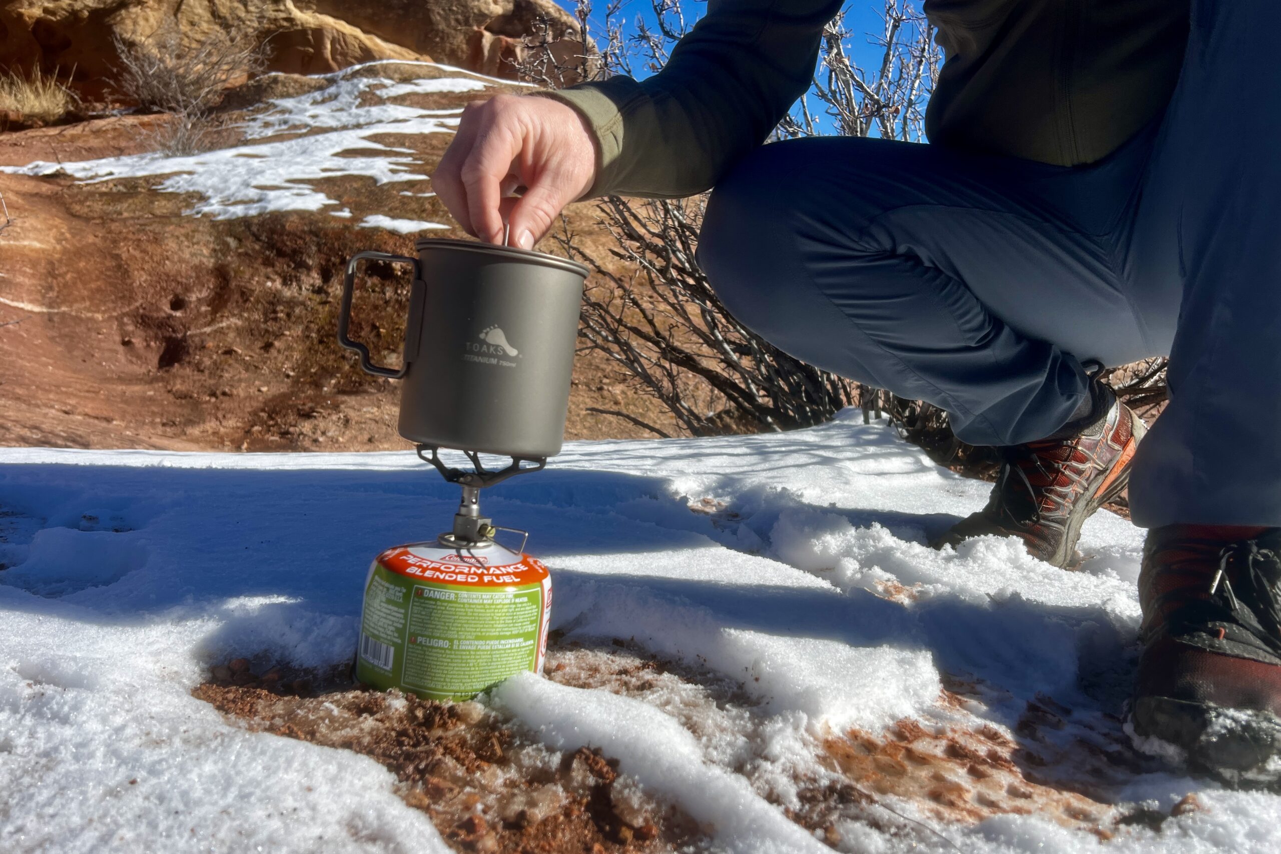 A hiker crouches next to a backpacking pot cooking on a stove and begins to remove the lid. Desert and snow background.