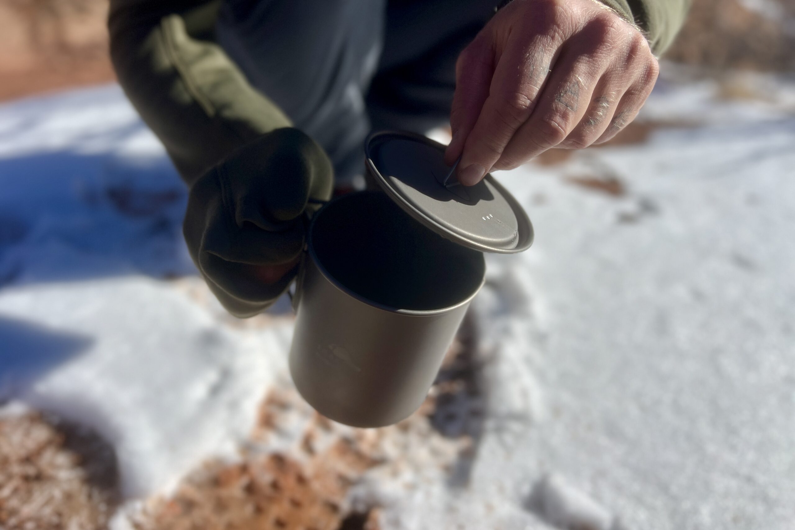 A hiker pulls the lid from a titanium backpacking pot against a snowy background.