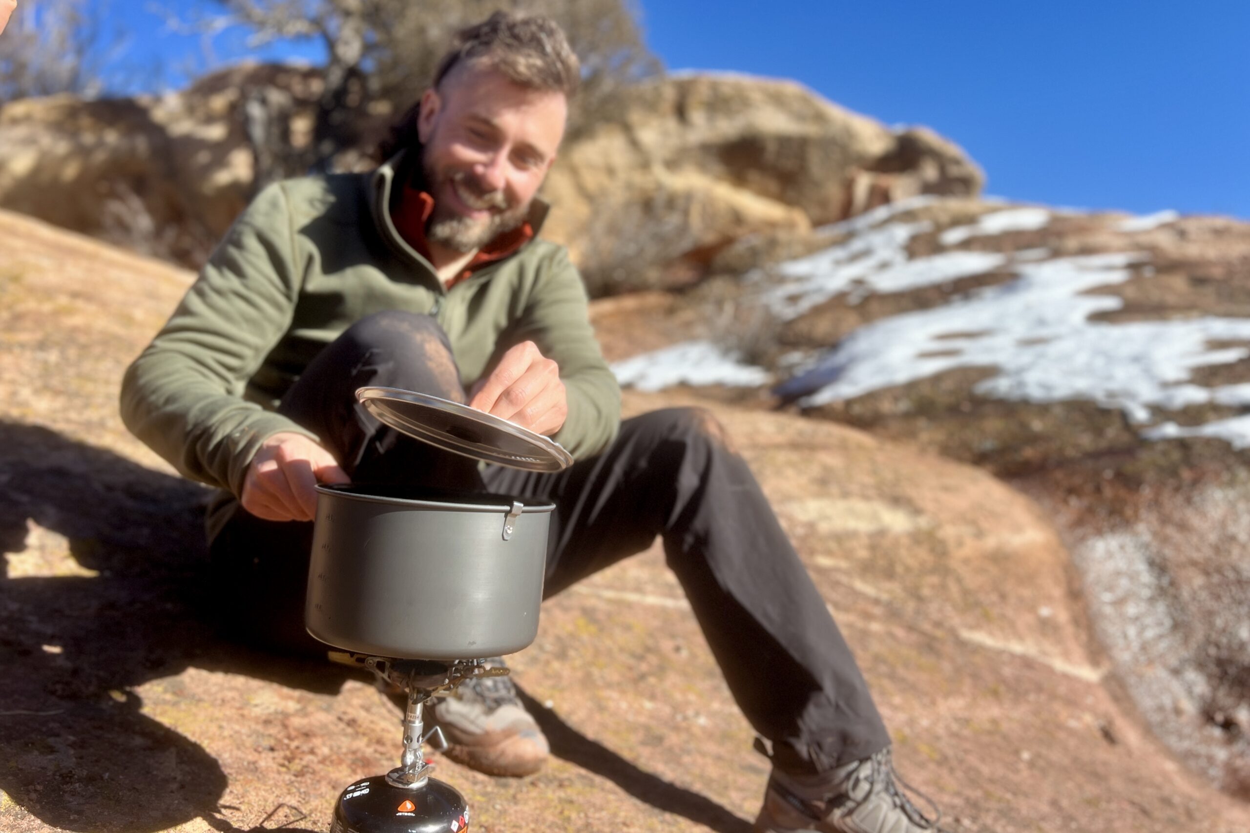 A hiker sits on the ground and lifts a pot's lid while cooking with it on a backpacking stove. Snowy desert background.
