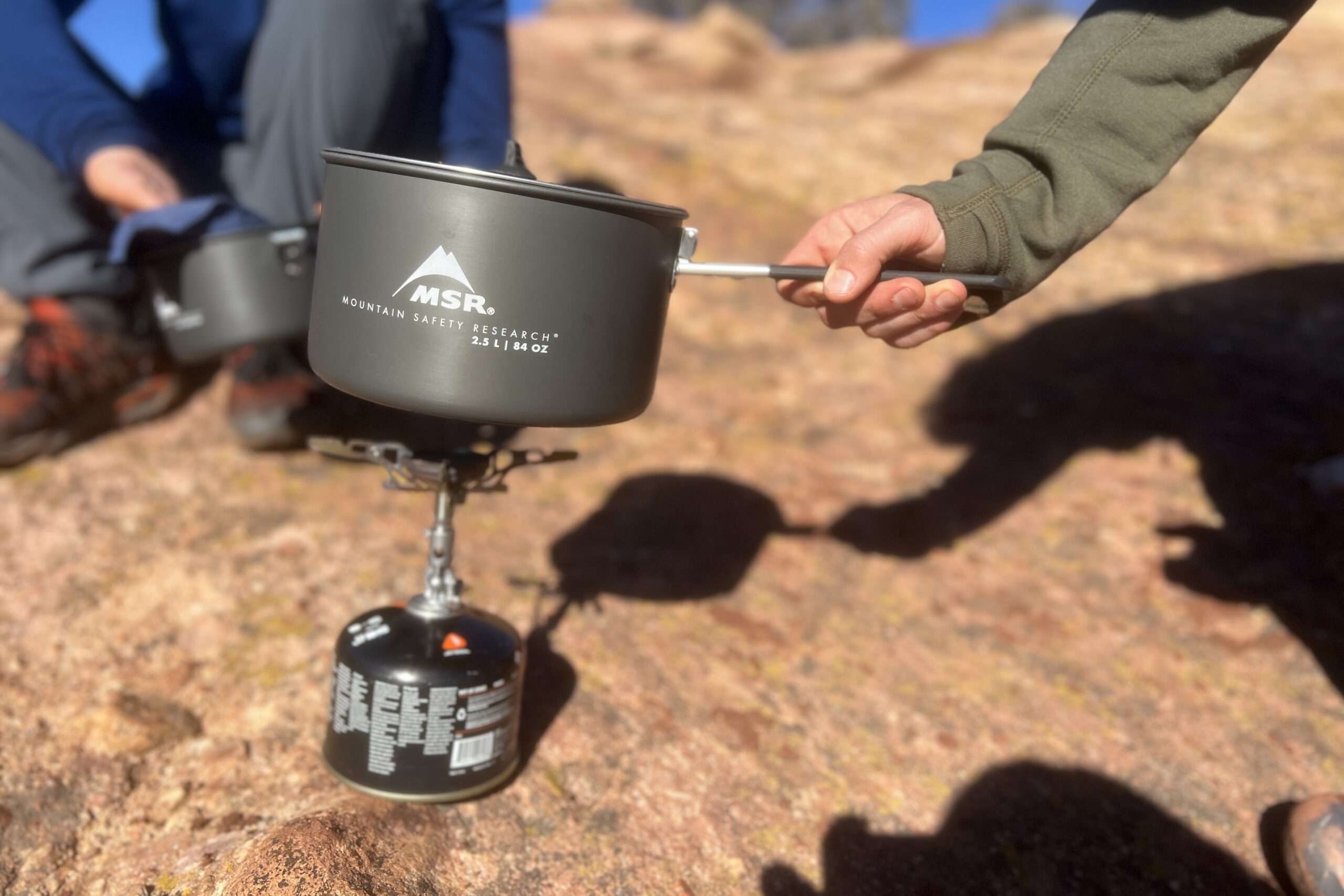 A hand places a pot on a backpacking stove while a hiker washes a pot in the background. Set in a rocky desert setting.