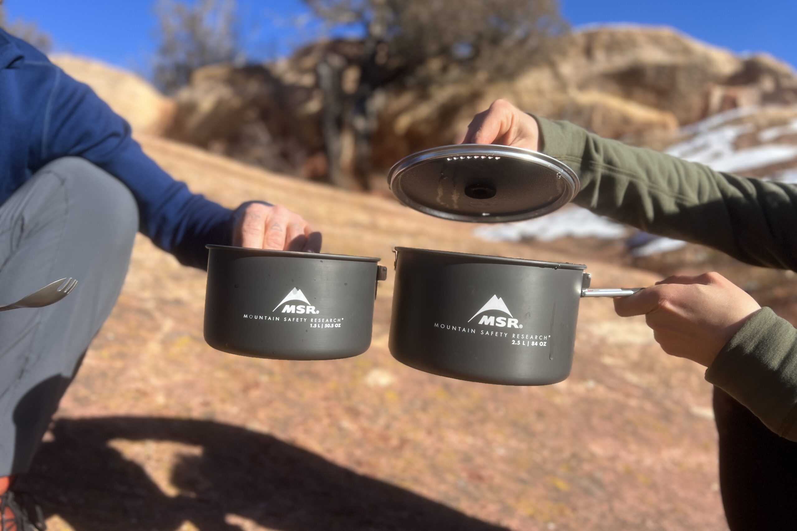 Two hikers show two different cook pots and a lid against a desert background.