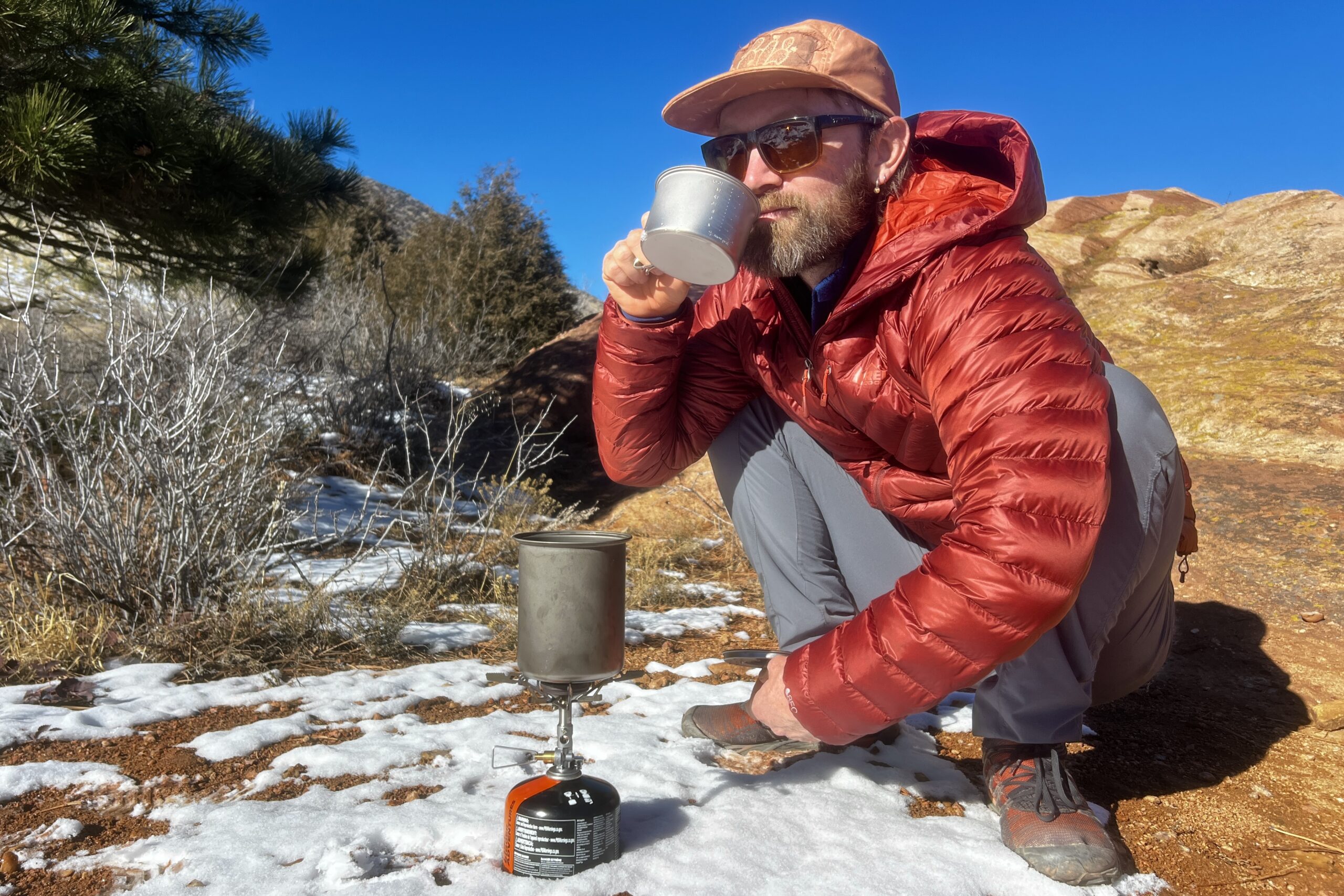 A hiker squats next to a camping cook set and sips from a small cup against a desert background.