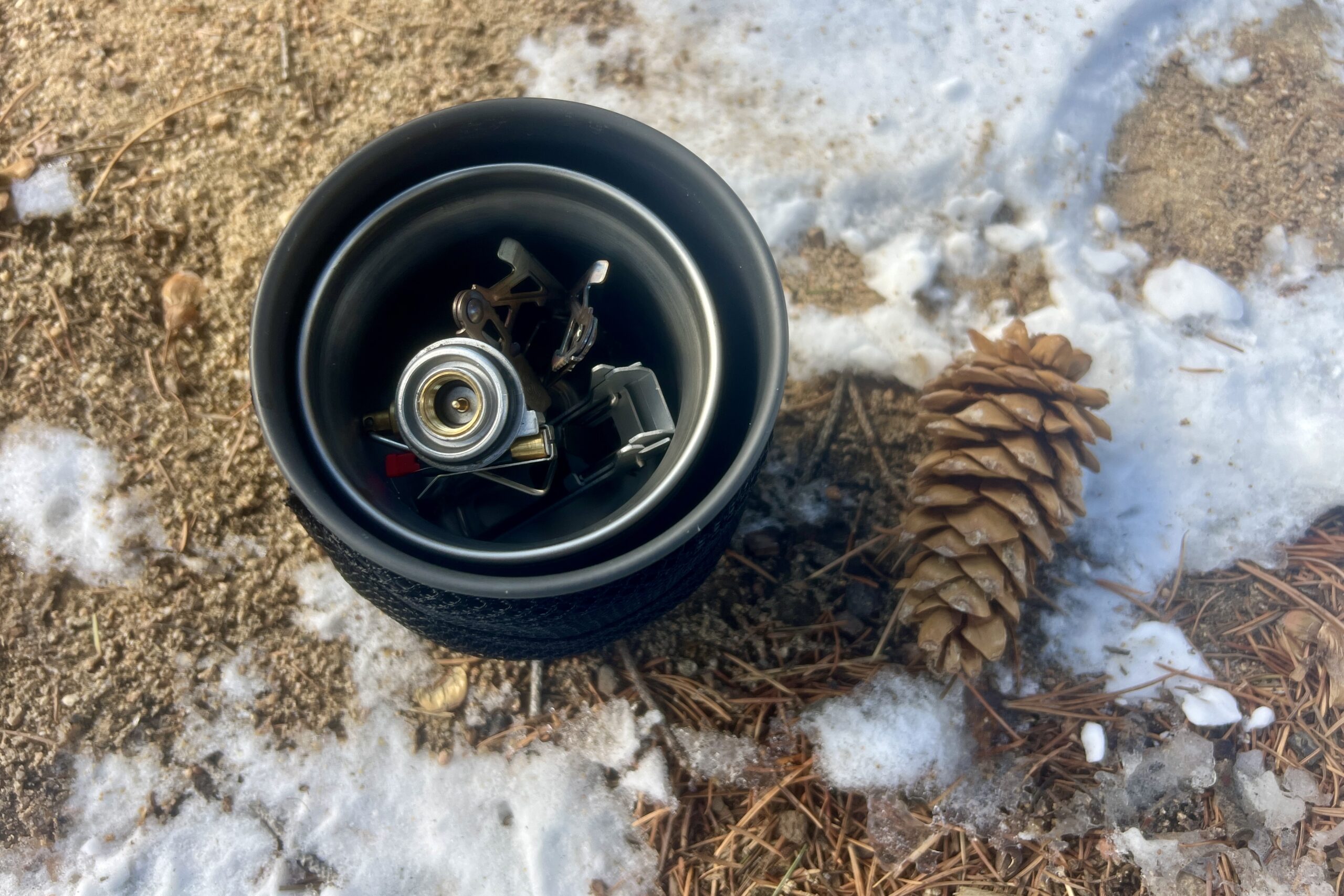 A top view of the thermostack combo's components nested together with a backpacking stove inside and a pine cone next to it against sandy ground.