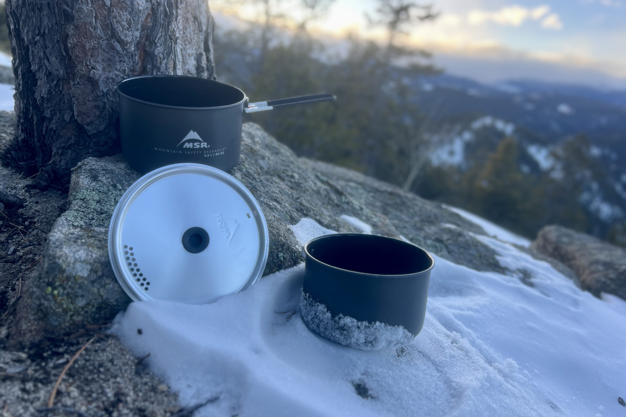 Two cook pots and a lid sit next to a tree with snow on the ground and a mountain sunset view.