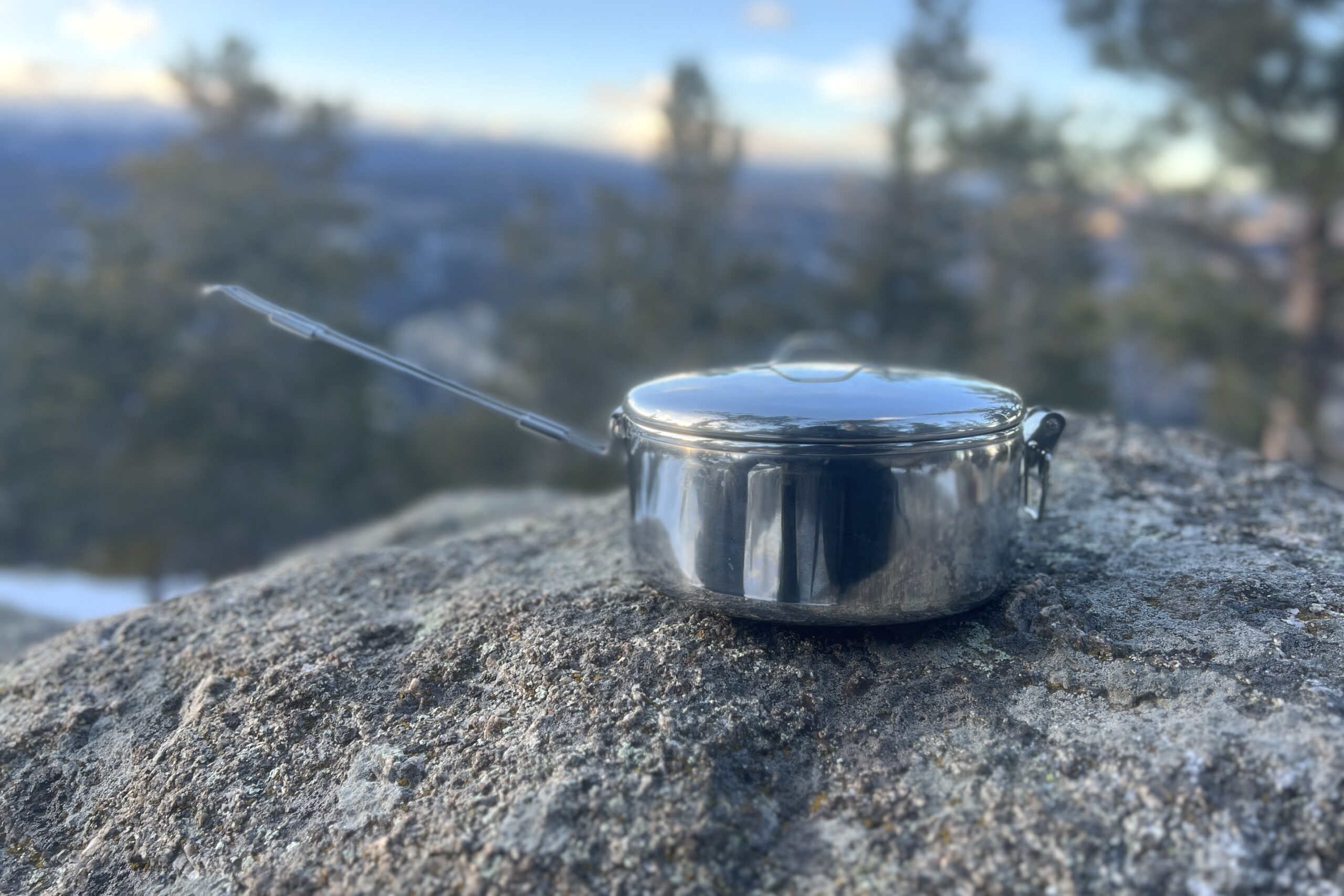 A steel backpacking pot with a foldable handle sitting on a rock