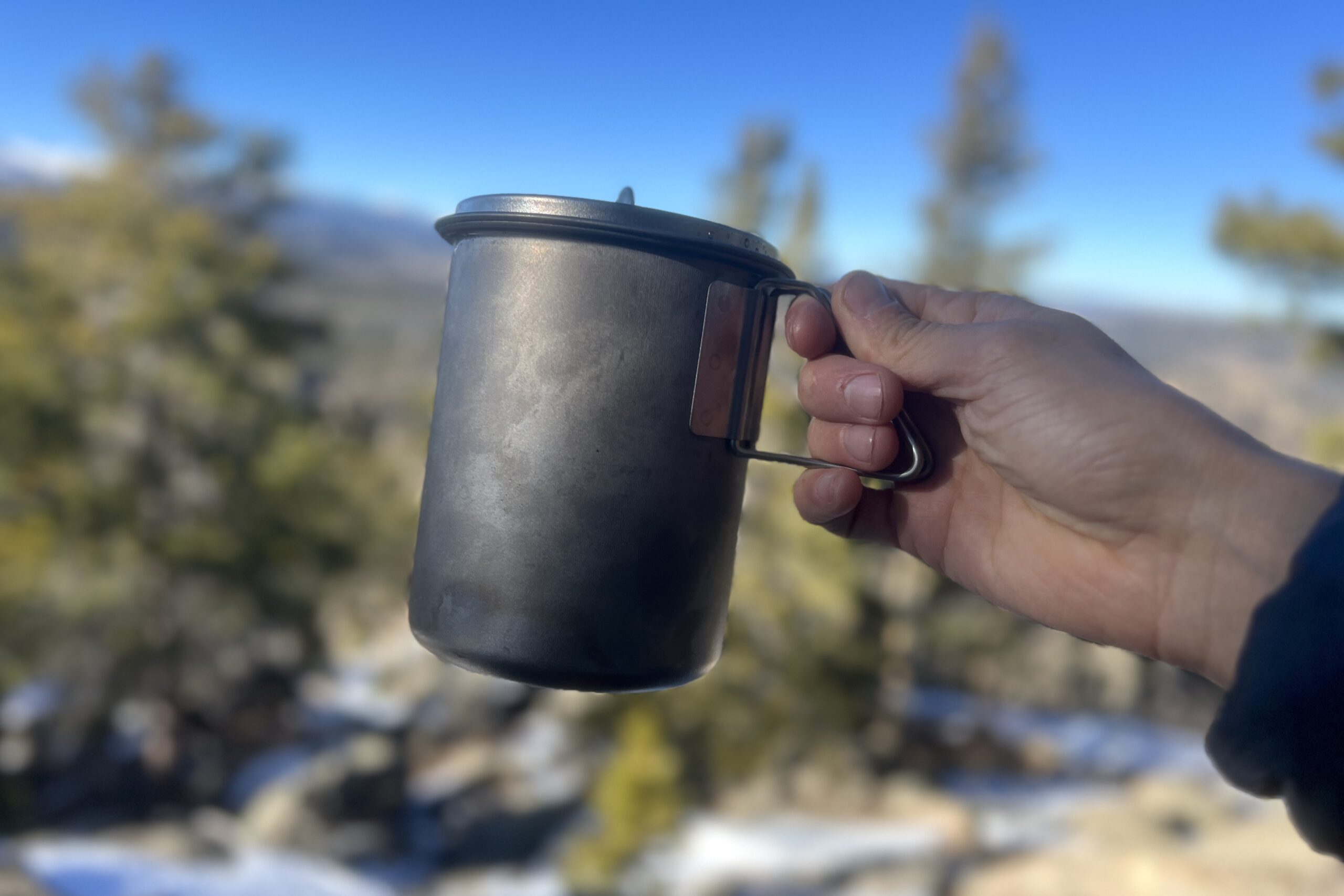 A hand holds a titanium cook pot with a lid.