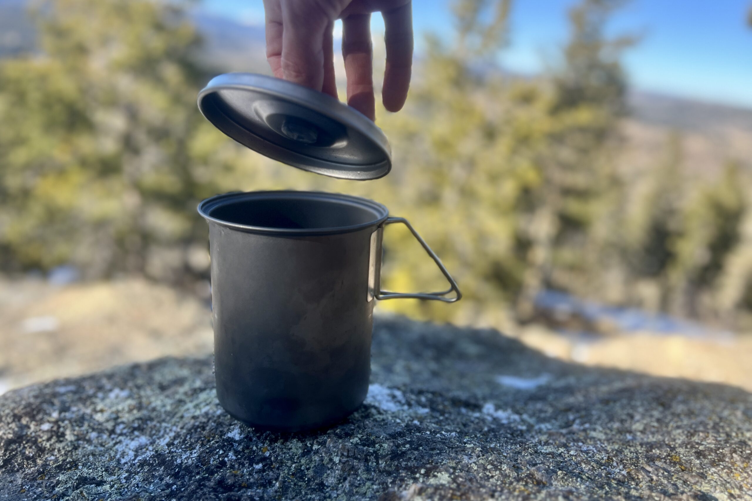 A hand removes the lid from a titanium cook pot.