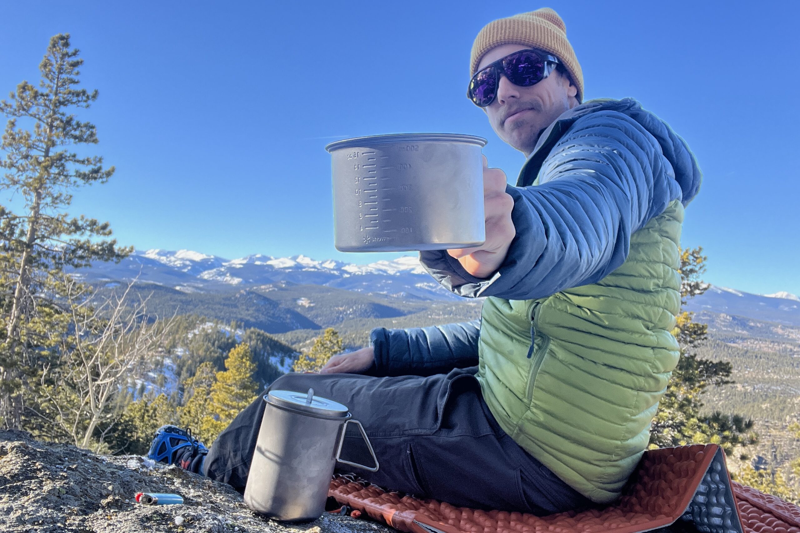 A hiker shows a titanium measuring cup to the camera with a blue sky and mountainous background.