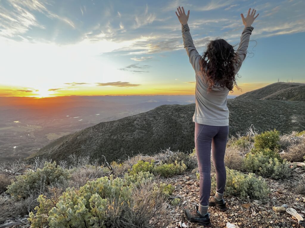 A woman stands before the sunset with arms outstretched over her head.
