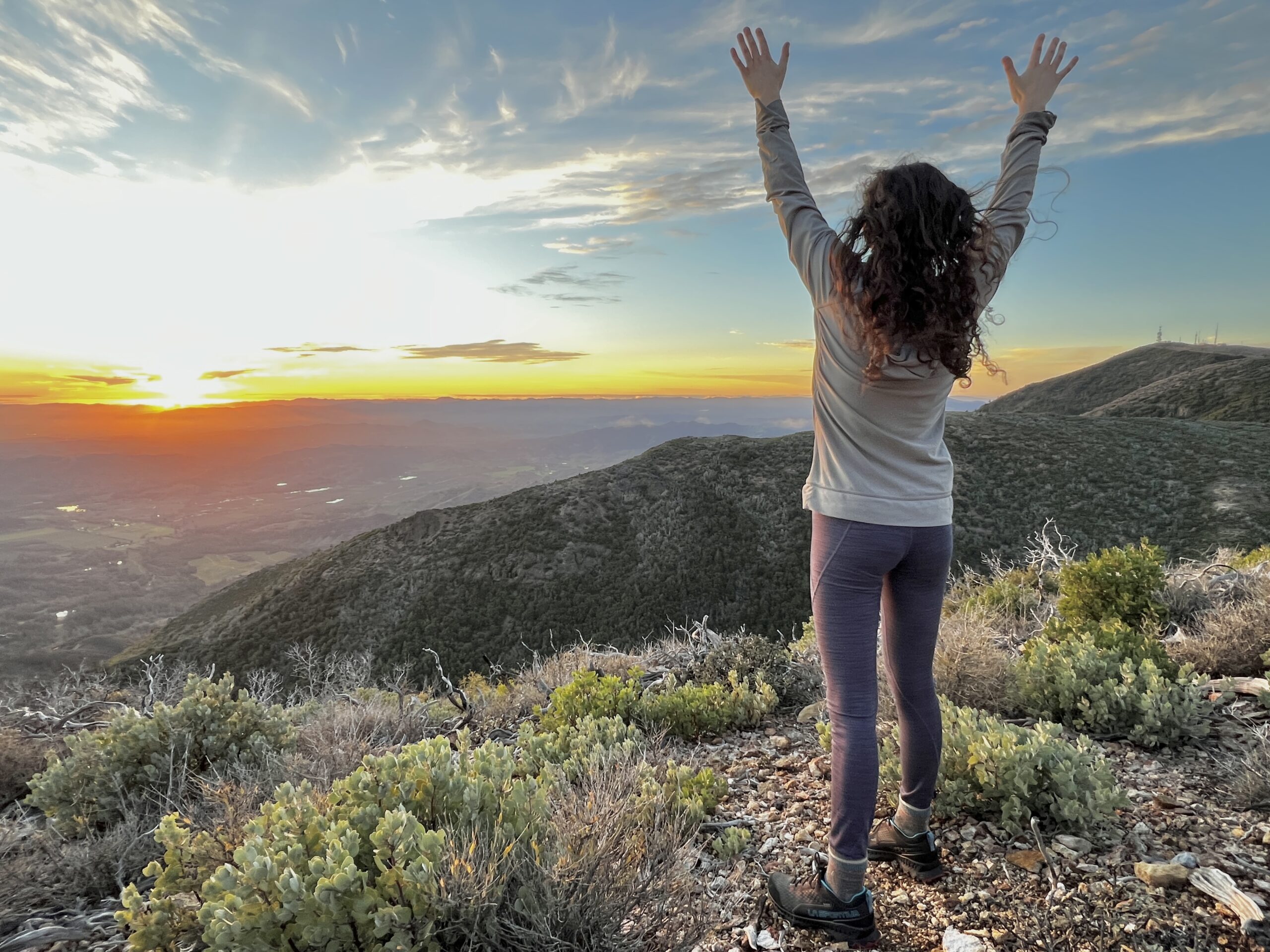 A woman stands before the sunset with arms outstretched over her head.