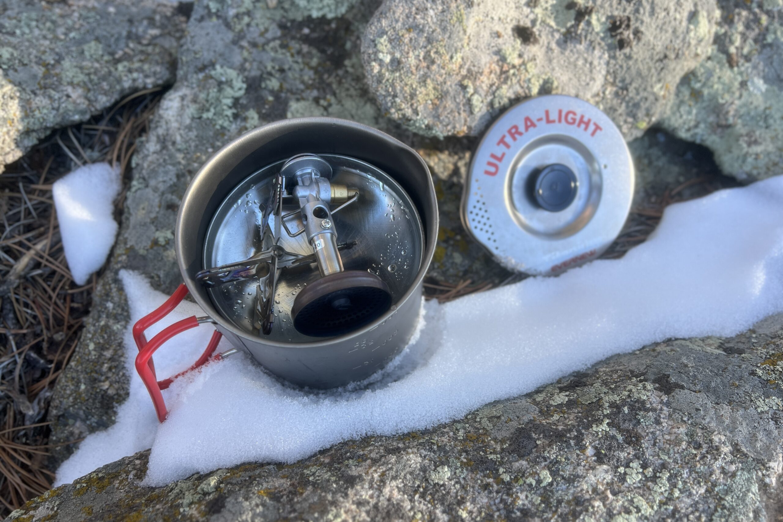 The Evernew pasta pot sitting on rock and snow with the lid nearby and a fuel canister and stove packed into the pot.