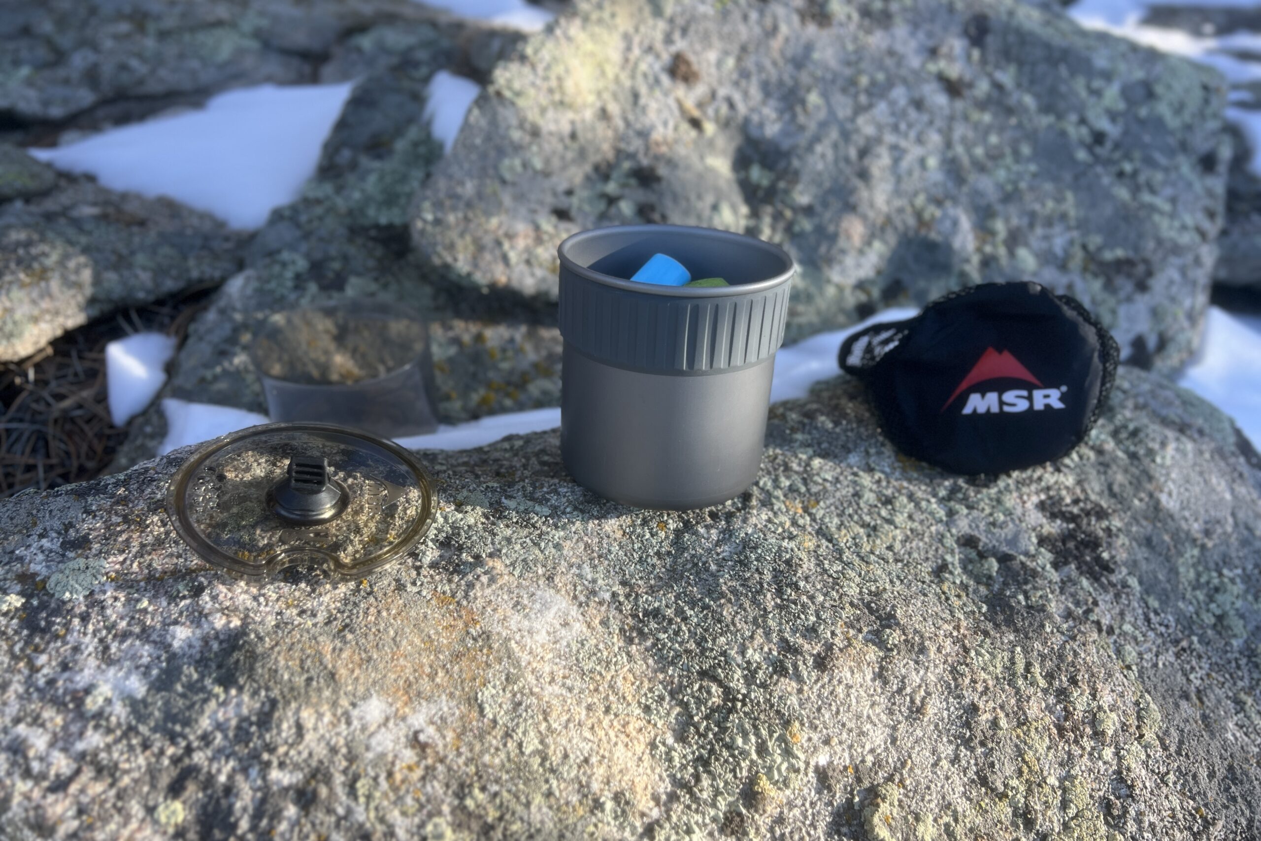 The lid, pot, and stuff sack of the msr trail mini solo sit next to each other on a rock.