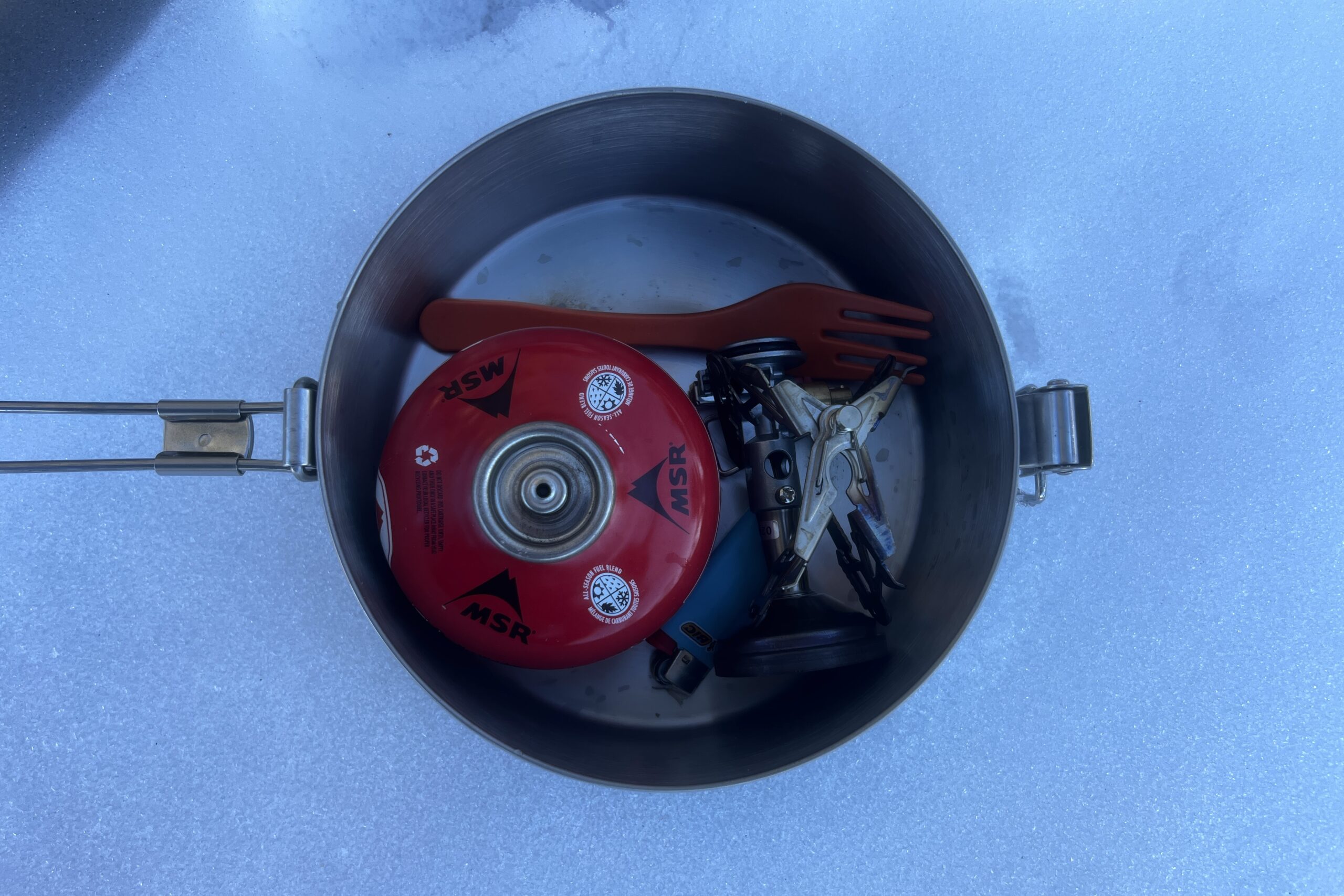 a top down view of fuel, a stove, fork, and lighter packed into a steel pot.