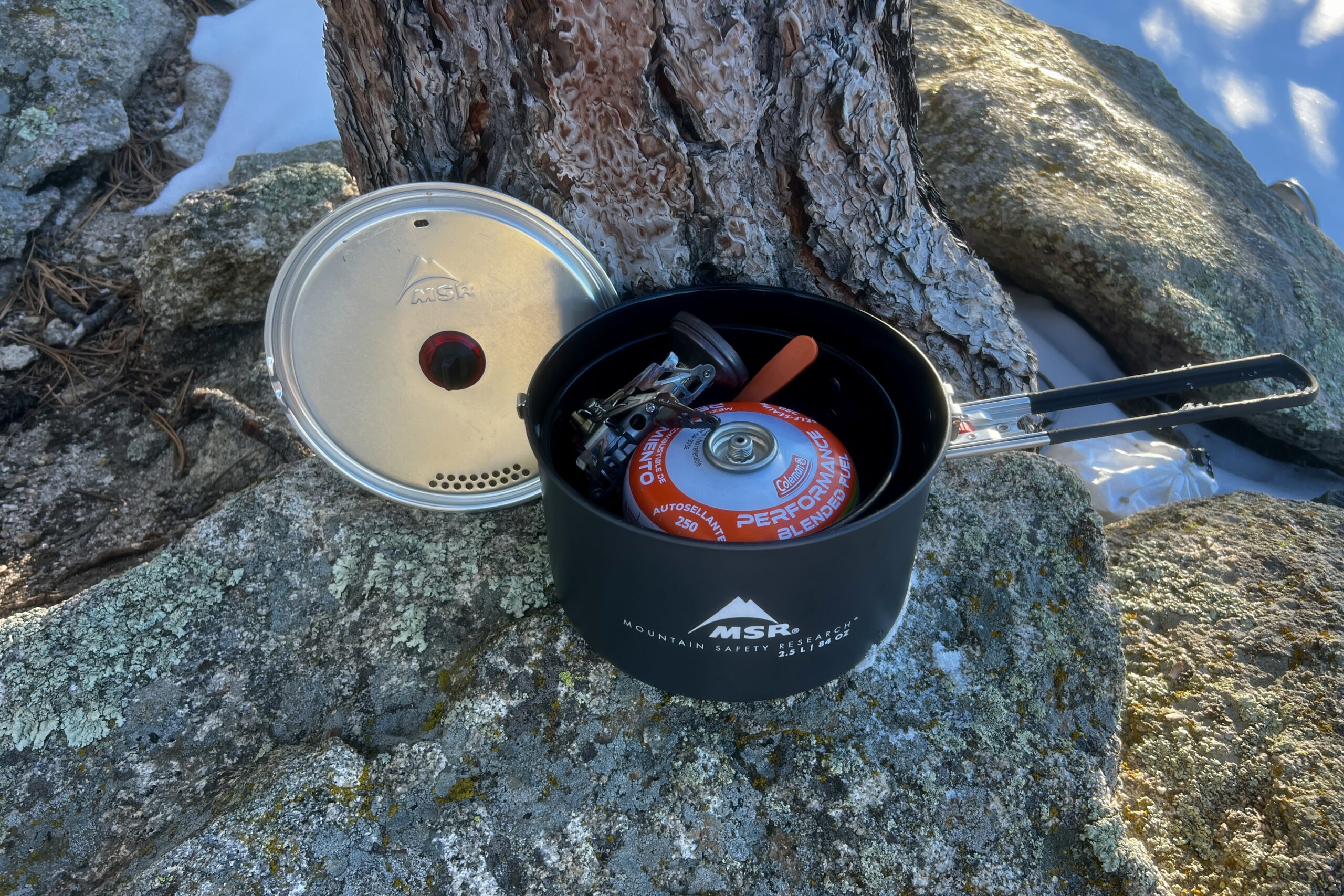 A pot and a lid sits next to a tree. A fuel canister, stove and other accessories are packed inside the pot.