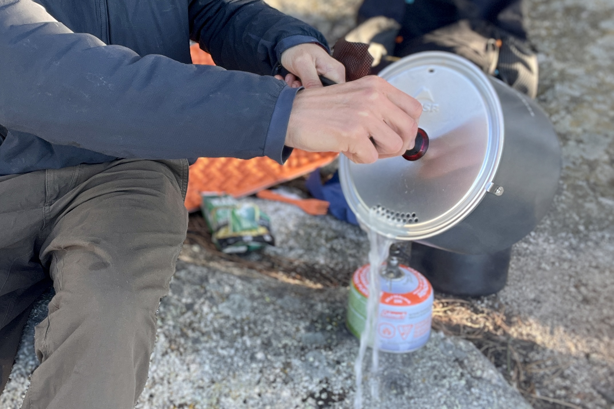 A hiker strains water out of a pot with backpacking gear strewn in the background.