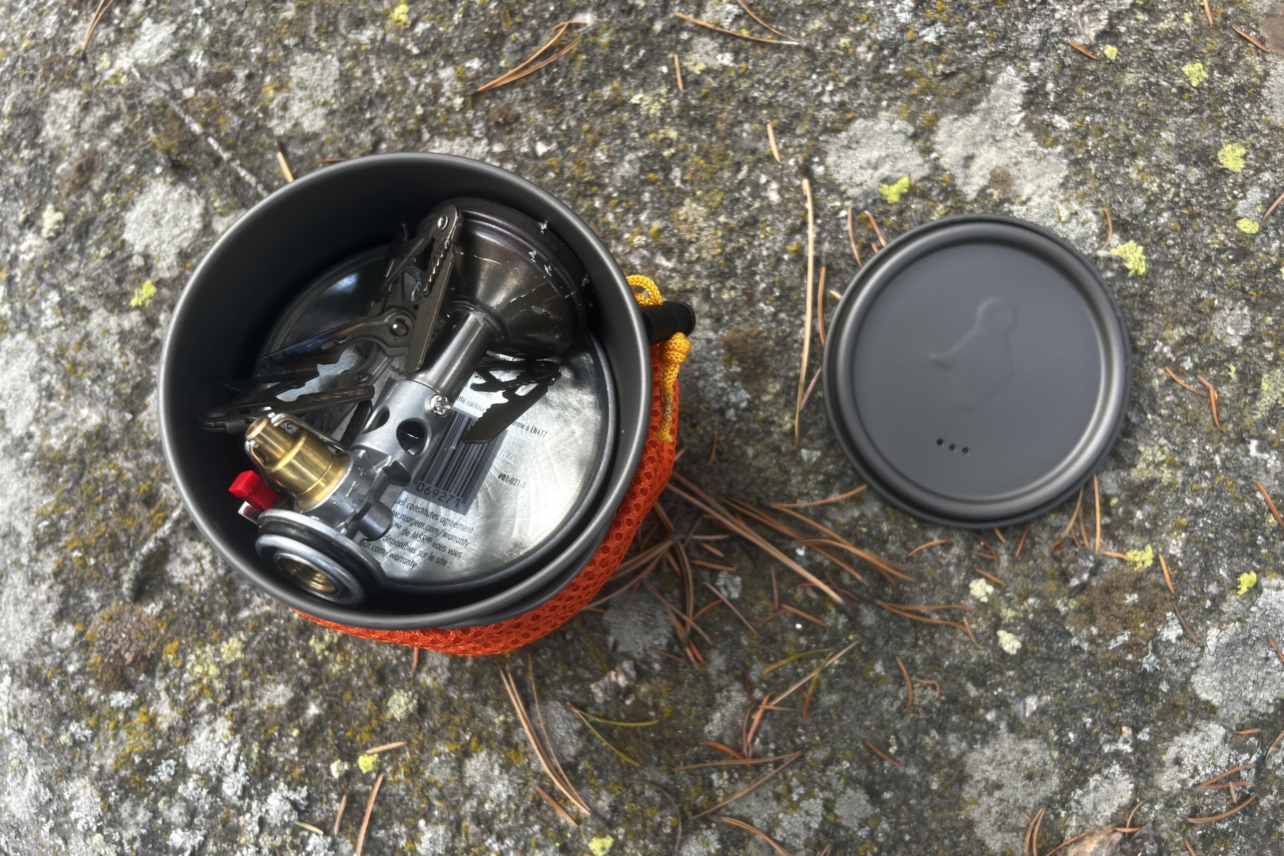The top down view of a stove and fuel packed into a backpacking pot with the lid next to it sitting on a rock.
