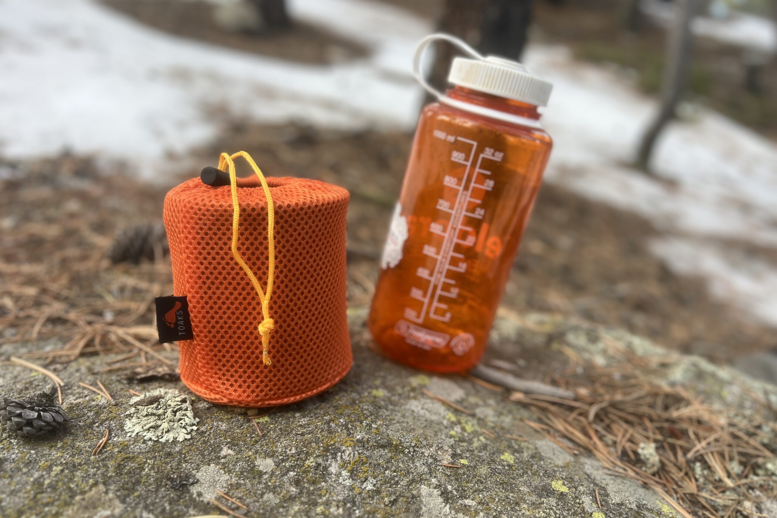 A cook set in an orange mesh stuff sack sits on a rock next to an orange nalgene bottle.
