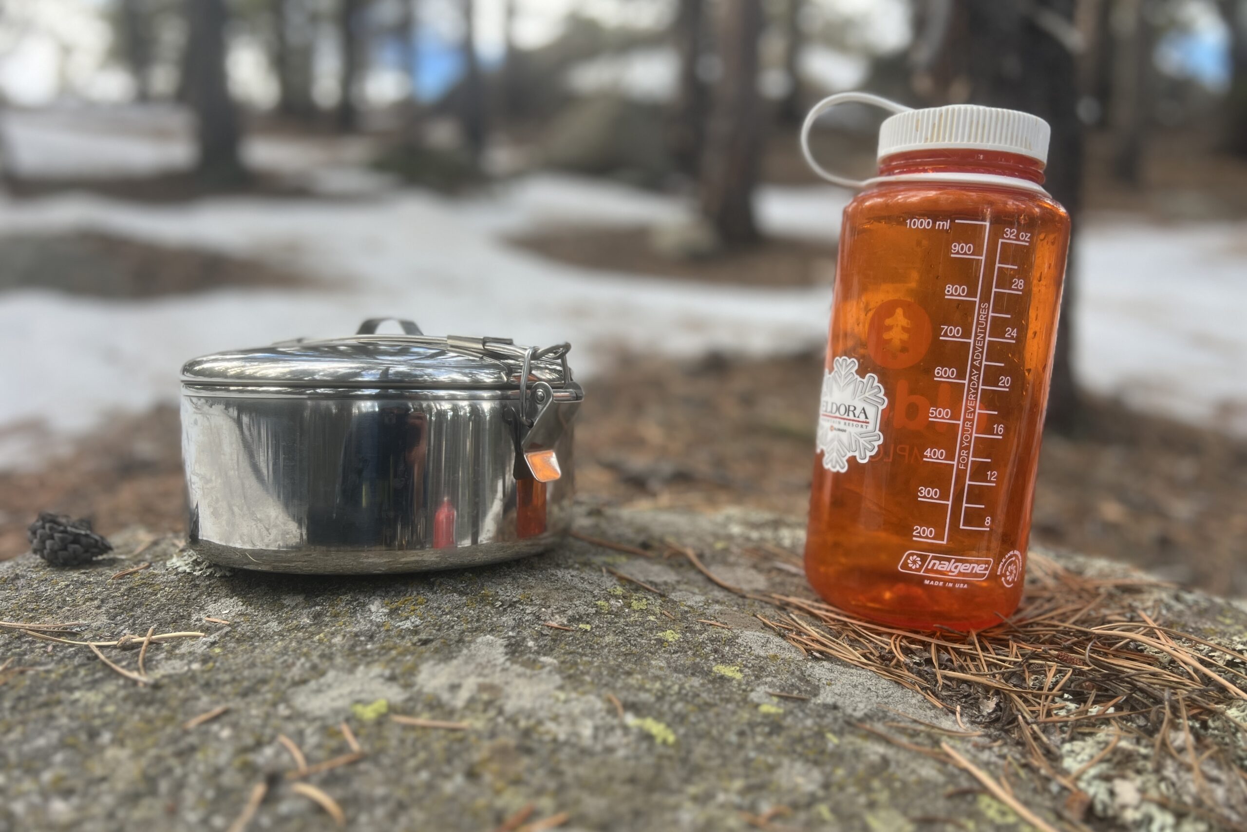 A steel backpacking cook set sits on a rock next to a standard nalgene bottle.