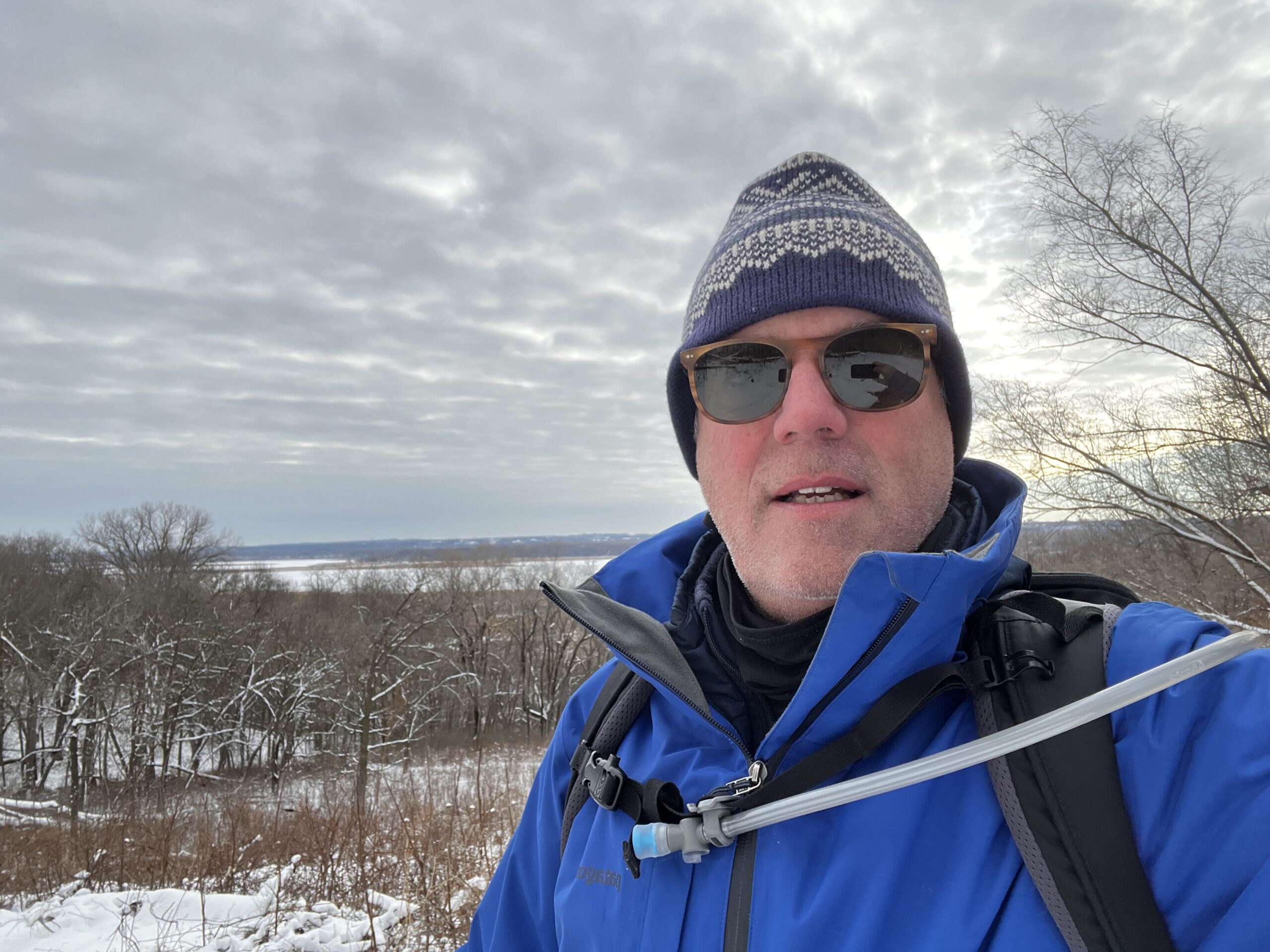 Image of a snowshoer wearing a winter hat and sunglasses with a snowy forest and lake in the background. A hydration hose is running out of his backpack and is attached to his sternum strap with a magnet.