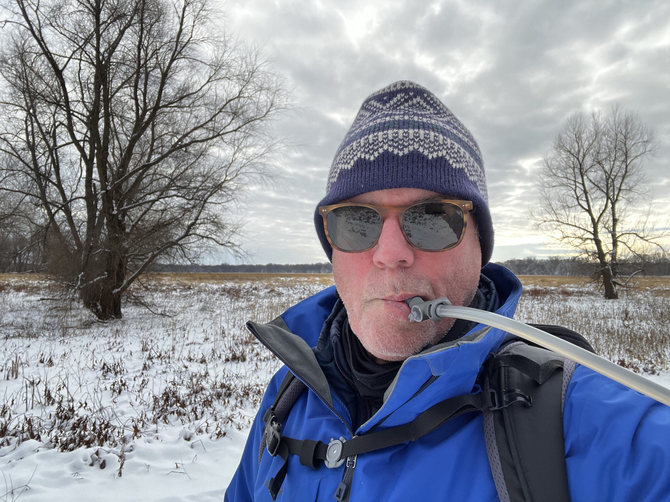 A snowshoer wearing a winter hat and sunglasses drinks from the HydraPak Contour's hydration hose. The background is a snowy field, with an overcast sky.