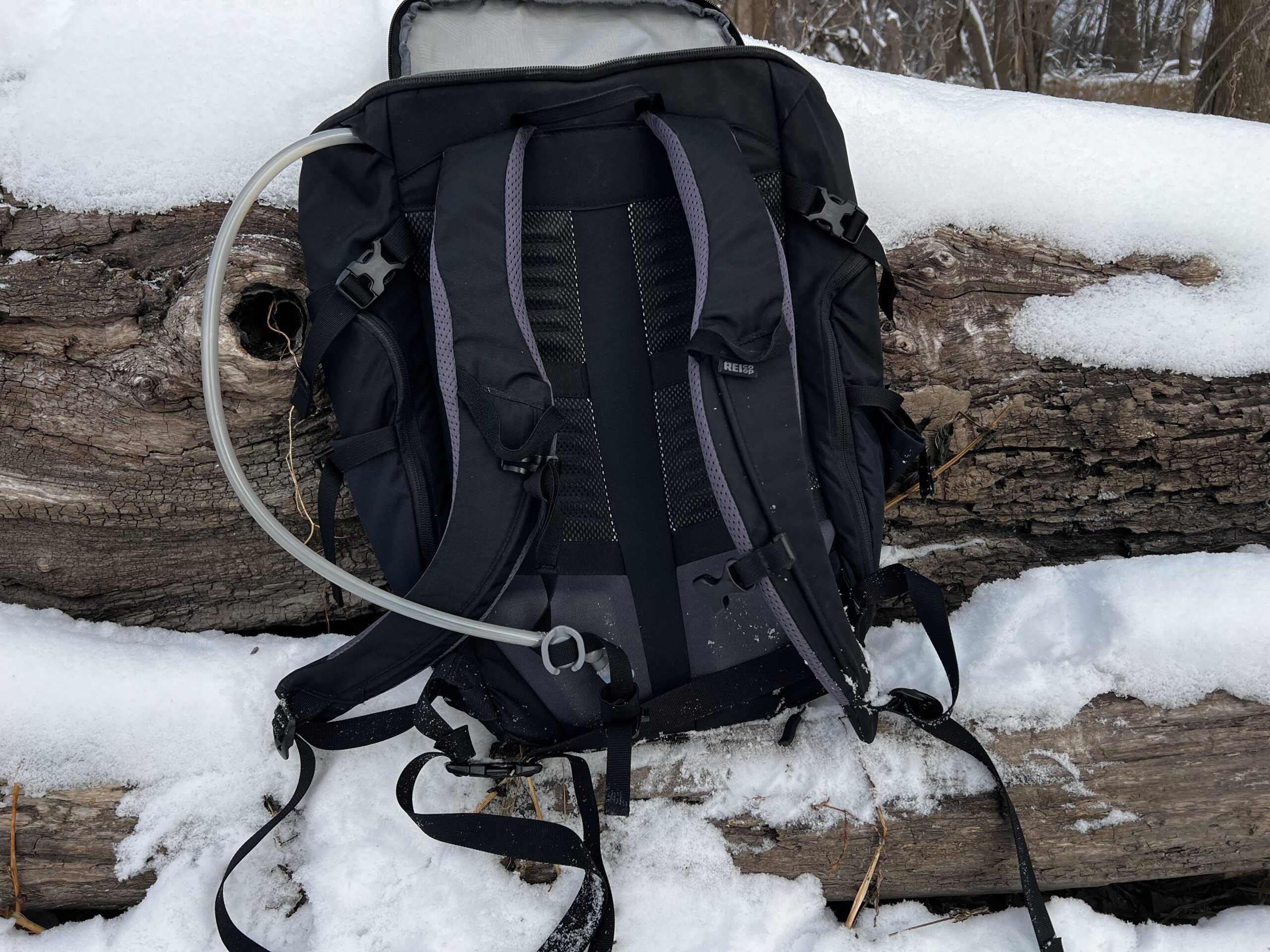 Close-up of a black backpack sitting on a snowy log. The HydraPak Contour's hydration hose is coming out of the top left side of the backpack. It is attached to the sternum strap with a magnet.