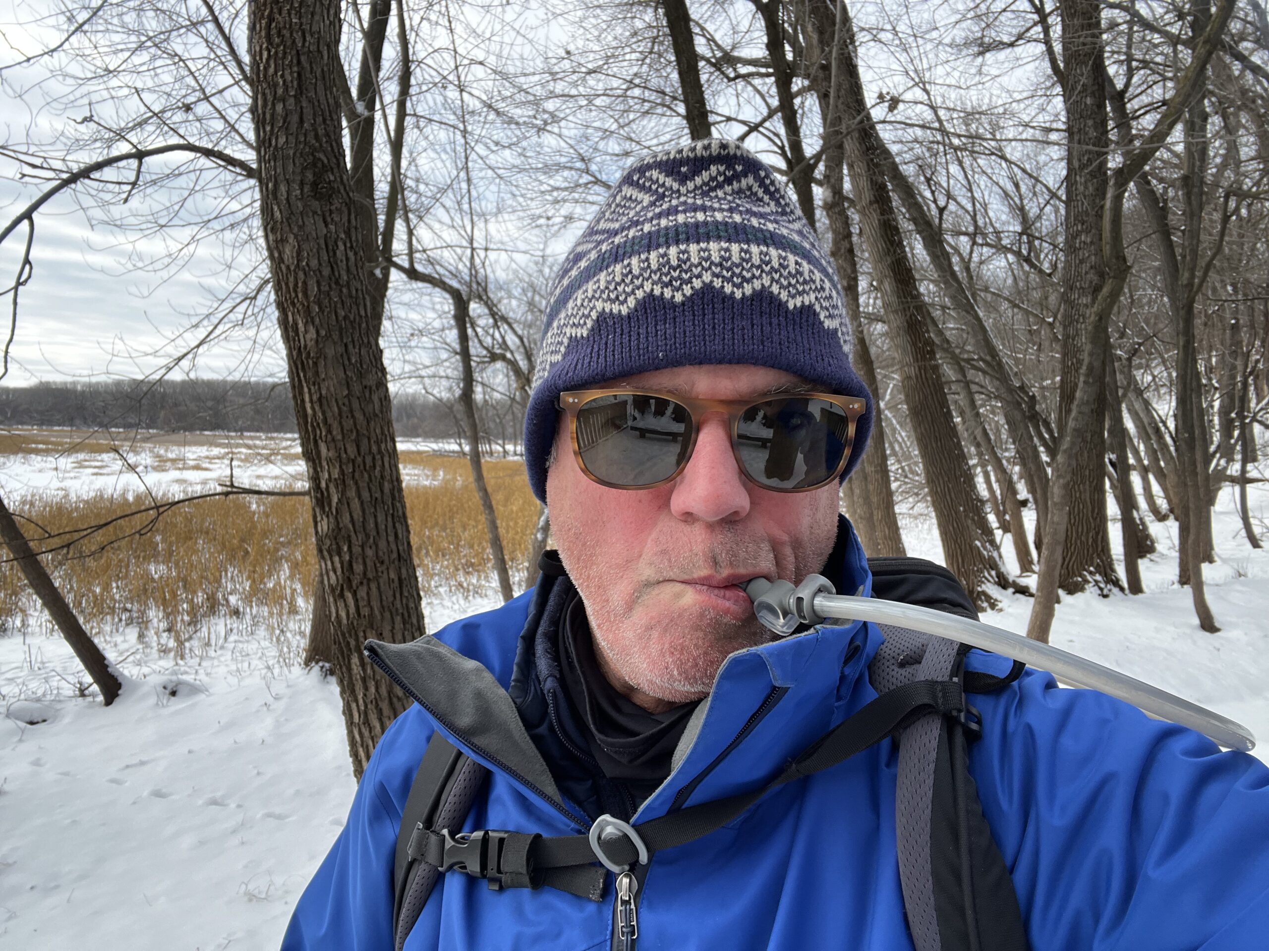 Close up of a snowshoer with a winter hat and sunglasses on. The background is snowy and lightly forested. The snowshoer is drinking from the HydraPak Contour's bite valve.
