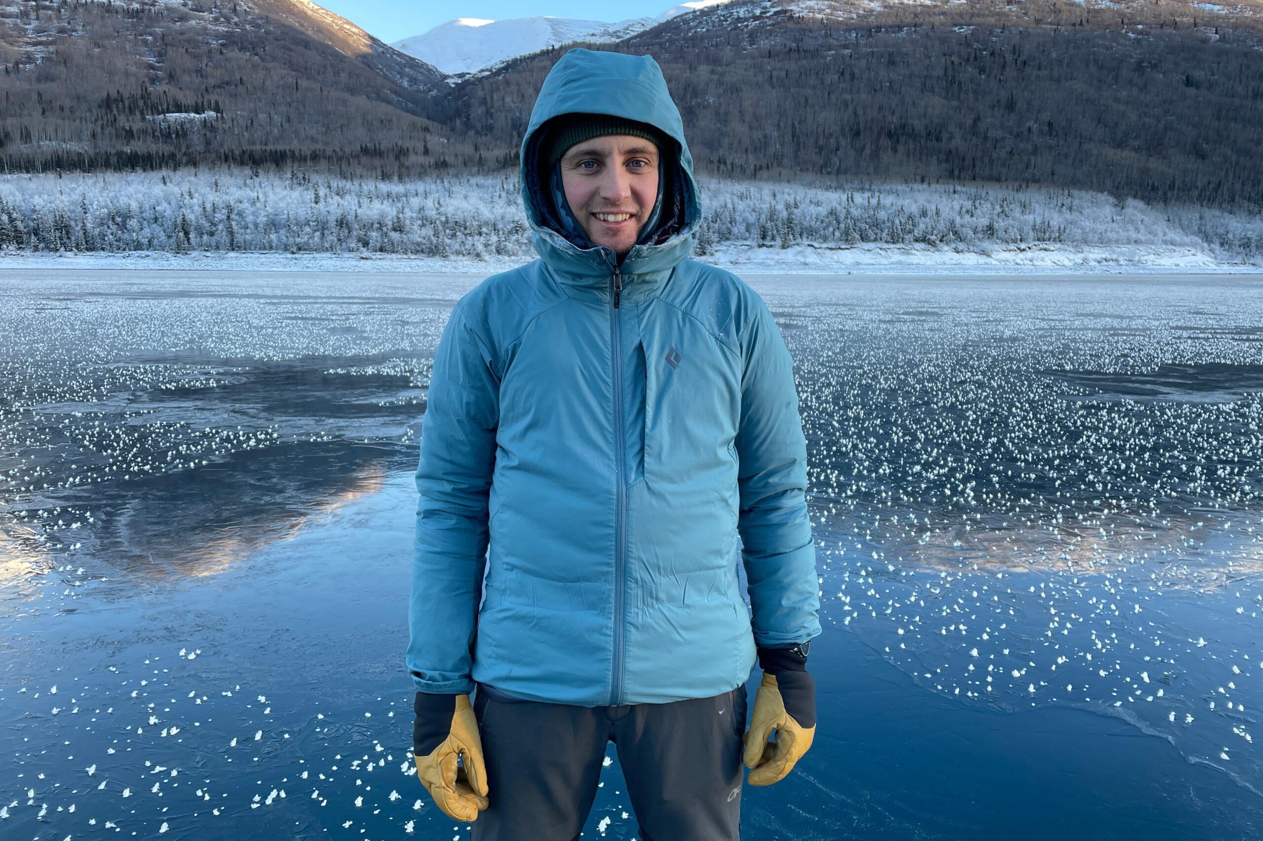 A man stands on a frozen lake with the hood of his jacket up.