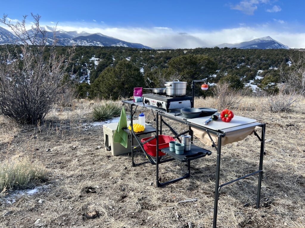 The GCI Master Cook Station stands outside with a mountain backdrop. Its shelves hold kitchen items and an outdoor stove. A cooler is on the ground next to it.