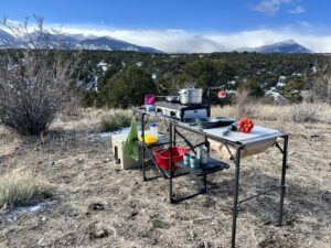 The GCI Master Cook Station stands outside with a mountain backdrop. Its shelves hold kitchen items and an outdoor stove. A cooler is on the ground next to it.