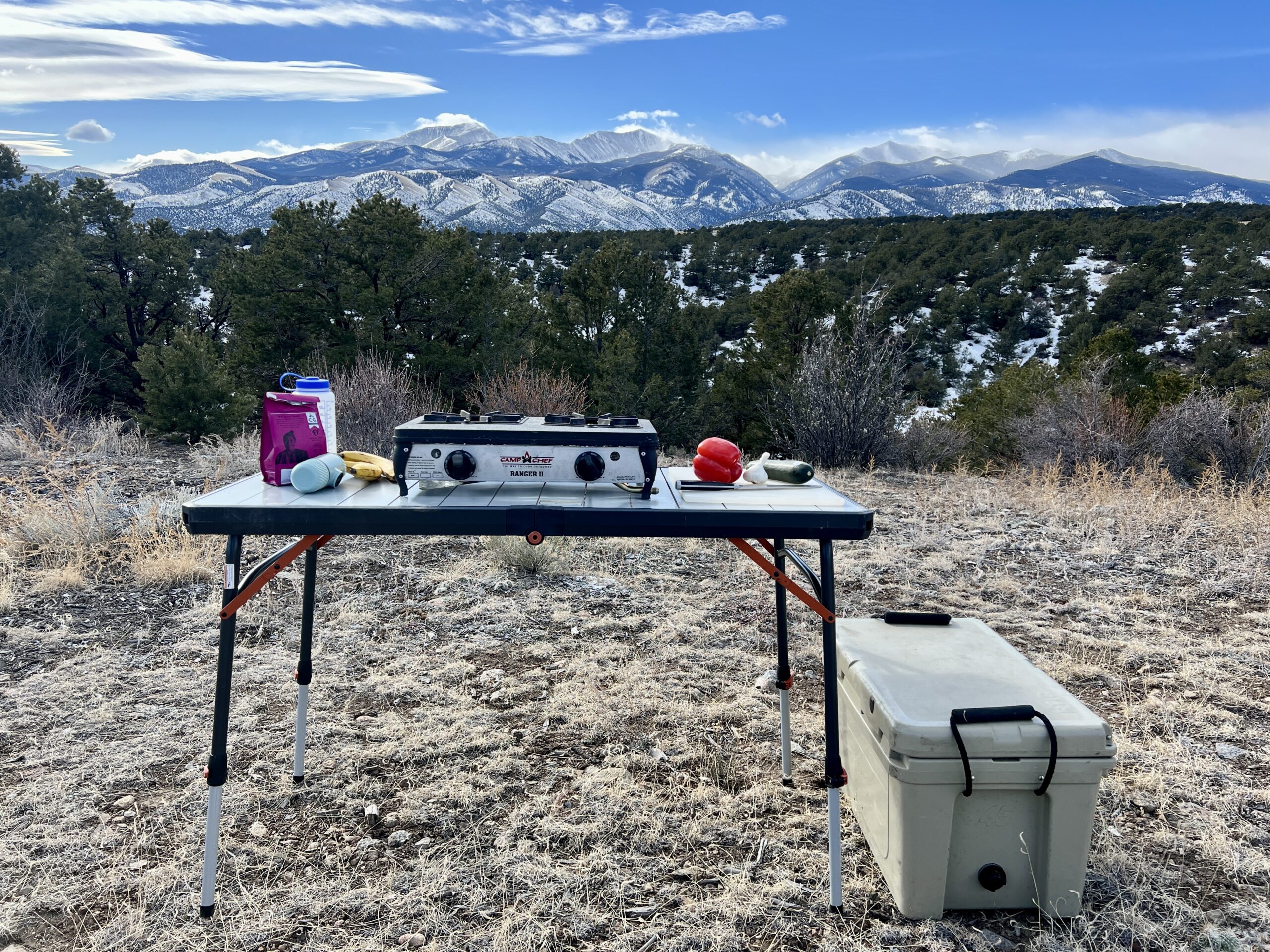 The REI Camp Prep Table has a stove, a red pepper and zucchini, a pink coffee bag, and bananas.