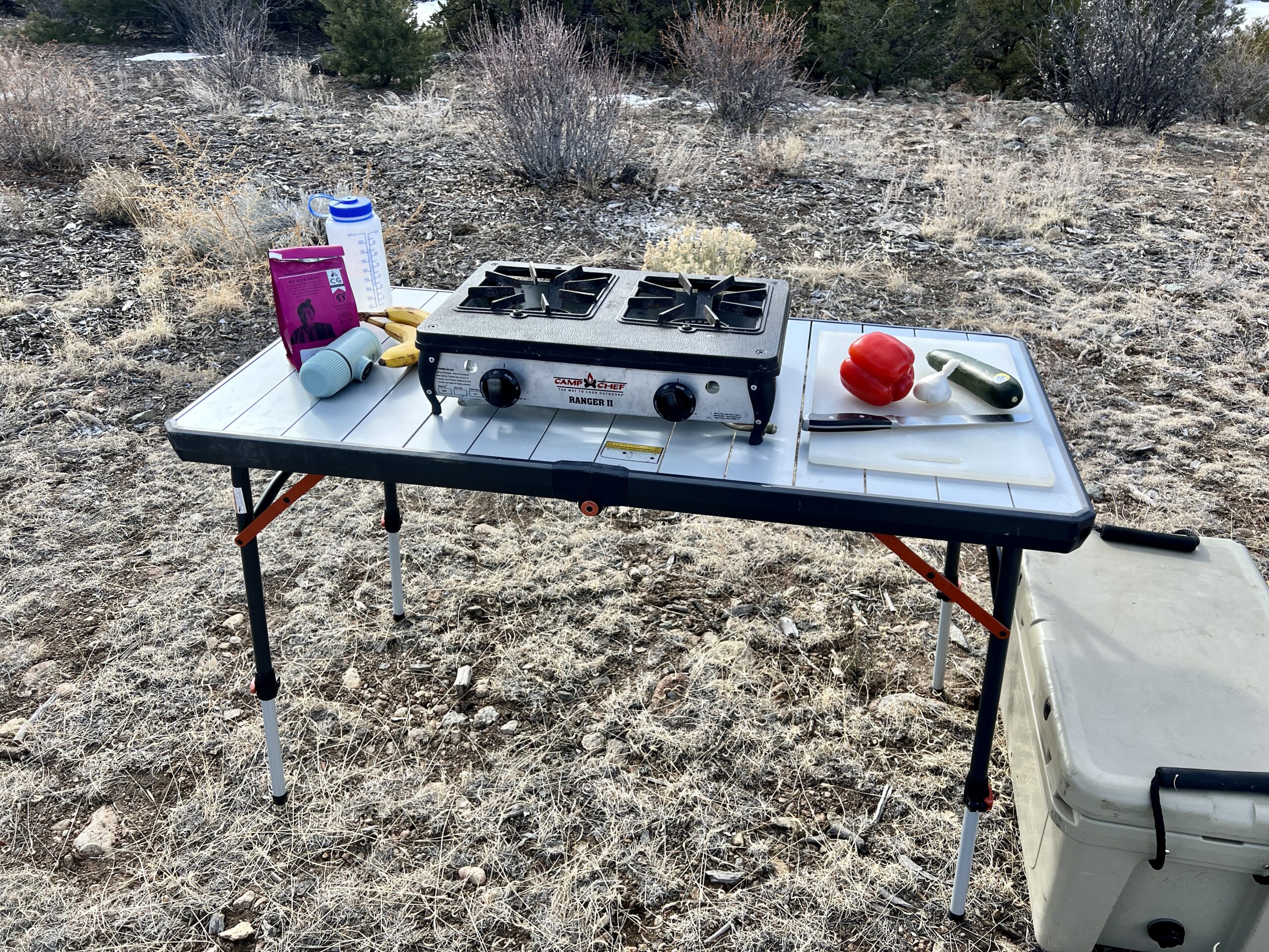 The REI Camp Prep Table has a two-burner stove, red pepper, zucchini, cutting board, knife, and garlic. A nalgene, pink coffee bag, bananas, and aeropress are on the left side.