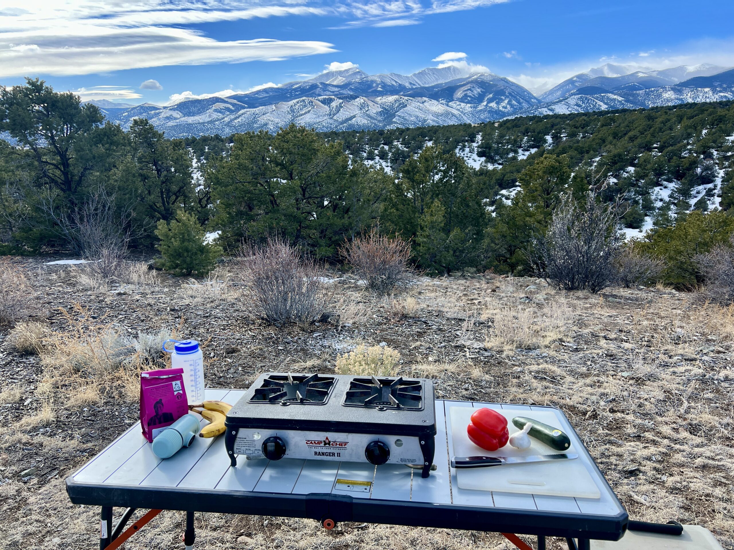 The REI Camp Prep Table has a two-burner stove, a cutting board with veggies, and a knife on top of it. On the left side, it has bananas, coffee, Nalgene, and aeropress.