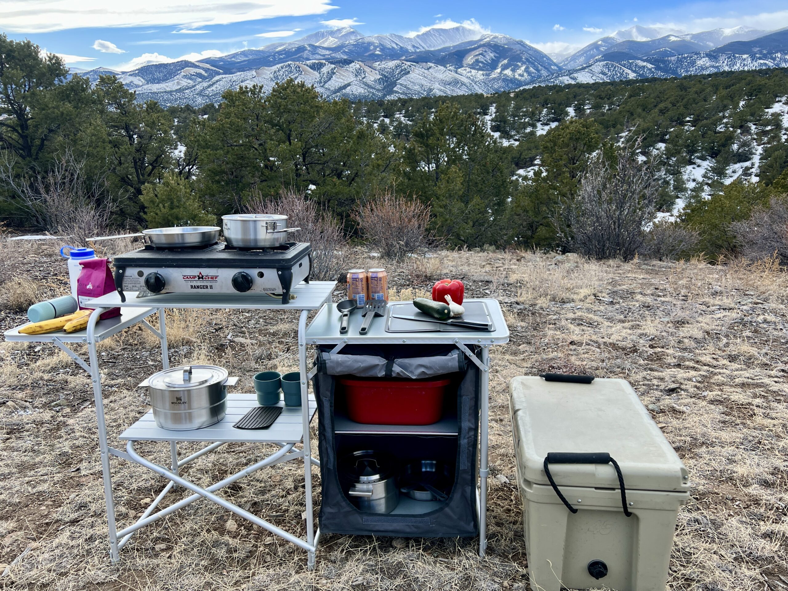 The MSG Deluxe Roll Top Kitchen stands on scrub ground with a mountain backdrop. It has kitchen cookware stored on its shelves and in its storage compartment. A cooler sits next to it.