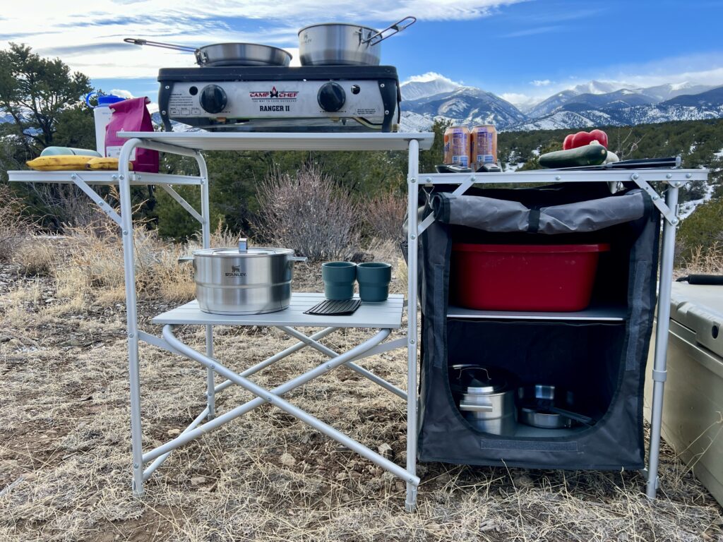 The Mountain Summit Gear Deluxe Roll Top Kitchen stands on scrub ground with a scenic mountain background. The table is loaded with cookware including pots and pans. An extra storage compartment is holding a red tub and more cookware.