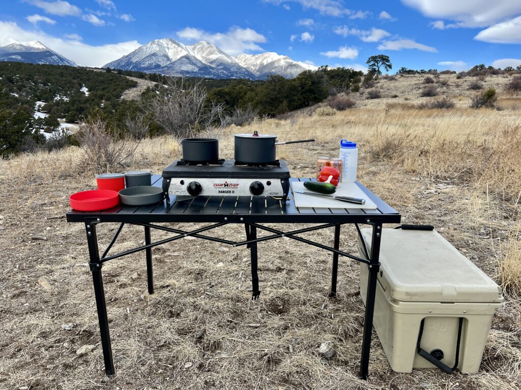 The Mountain Summit Gear Heavy Duty Roll Top Table stands on scrub ground with a scenic mountain backdrop. The table has red and gray cookware
