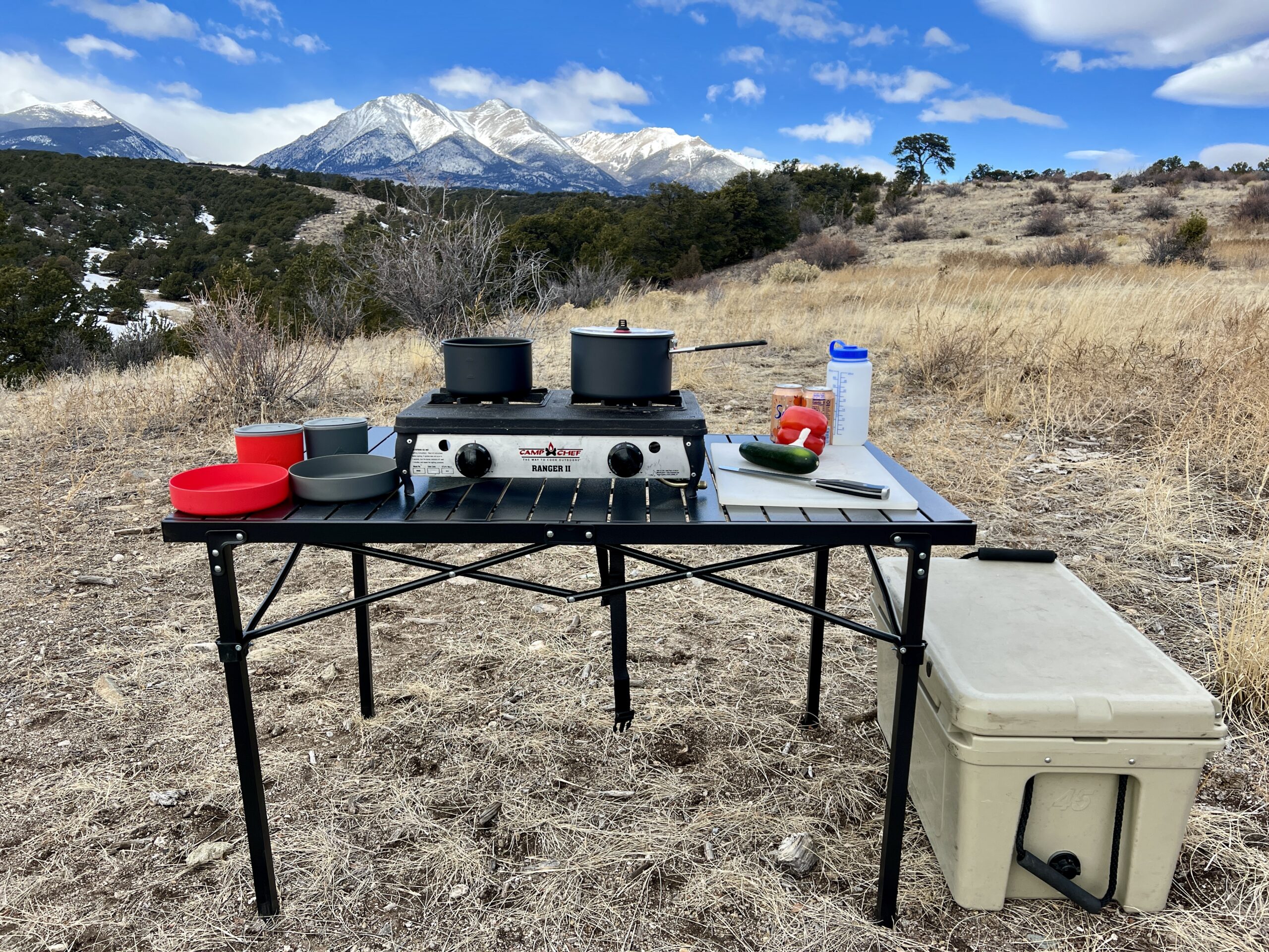 The Mountain Summit Gear Heavy Duty Roll Top Table stands on scrub ground with a scenic mountain backdrop. The table has red and gray cookware
