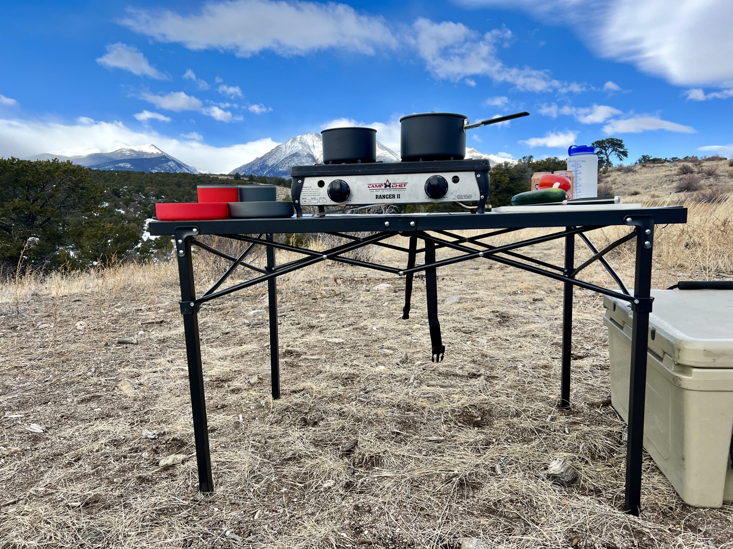 The Mountain Summit Gear Heavy Duty Roll Top Table stands on scrub ground with a scenic mountain view in the background. A two-burner stove, red and gray cookware, and some food items are on top. A beige cooler is sitting on the right side.