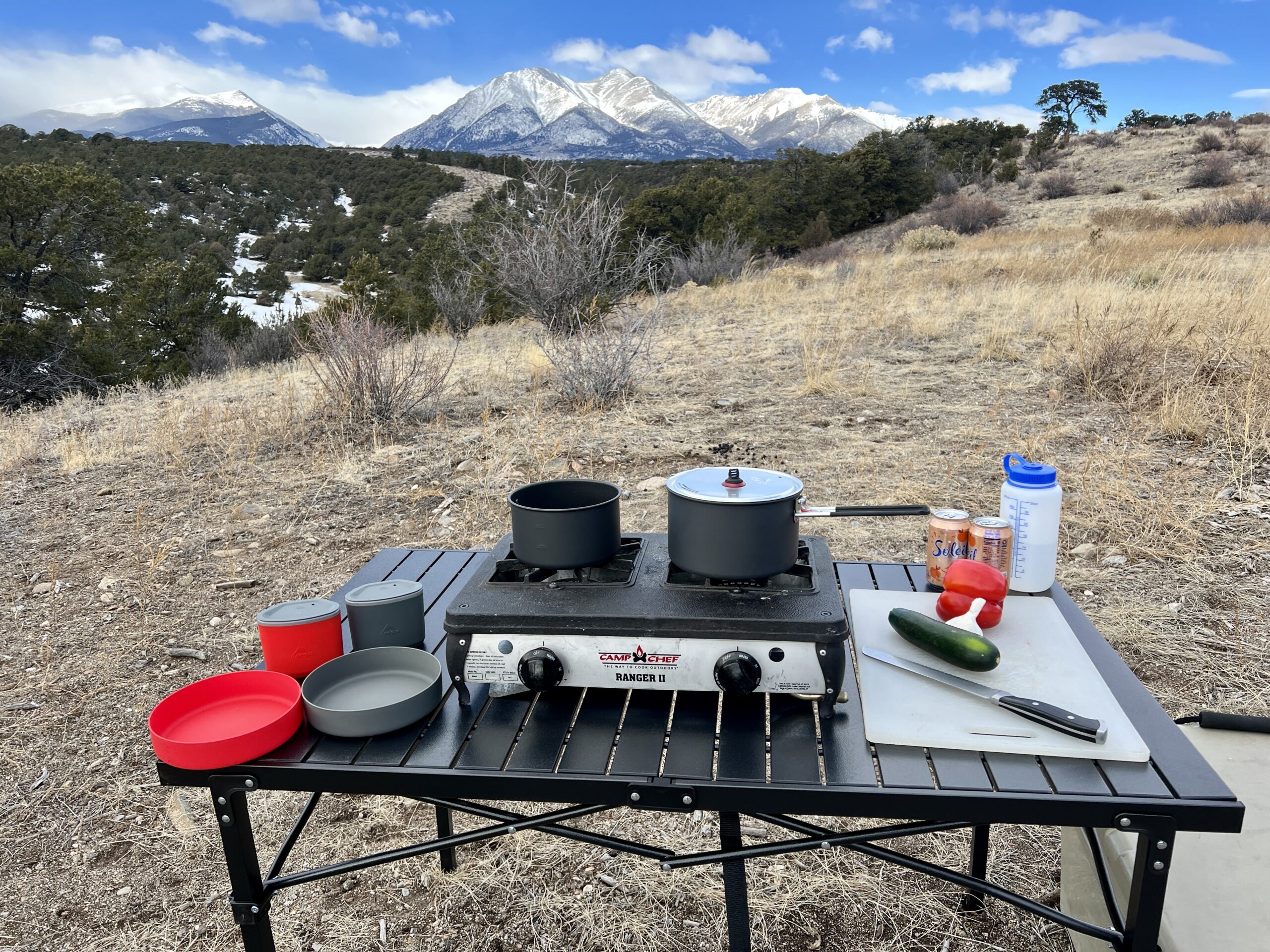 The Mountain Summit Gear Heavy Duty Roll Top Table stands on scrub ground with a scenic mountain view in the background. A two-burner stove, red and gray cookware, and some food items are on top. A beige cooler is sitting on the right side.