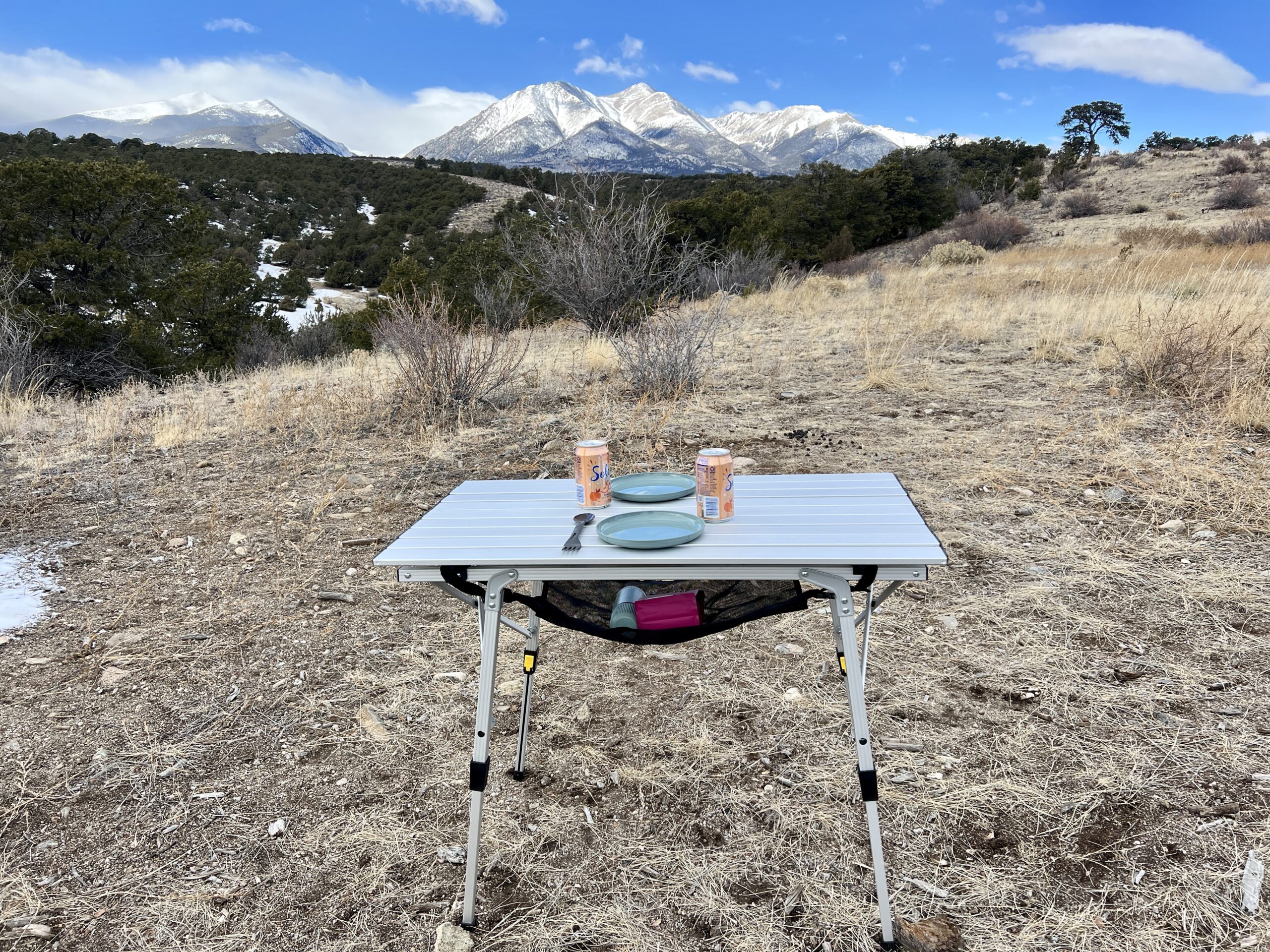 The Portal Quick Adjust Lightweight Roll-Up Table Roll-Top Table stands on scrub with a scenic mountain backdrop. Teal plates and cans of water are on the table. There is a pink coffee bag and a teal coffee maker under the table.