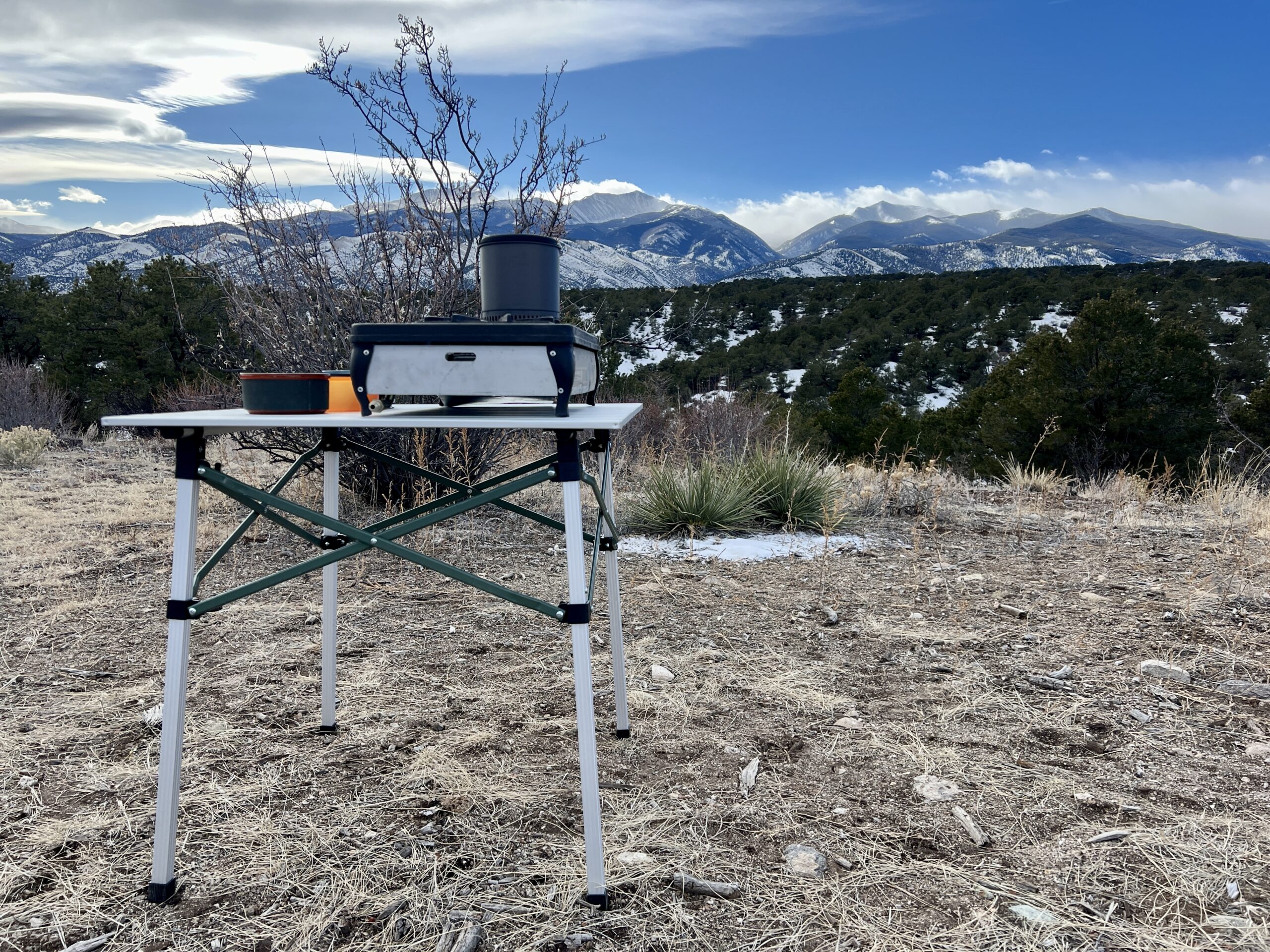 The REI Camp Roll Table is standing outside with a mountain backdrop. It has a stove and two plastic bowls on its top.