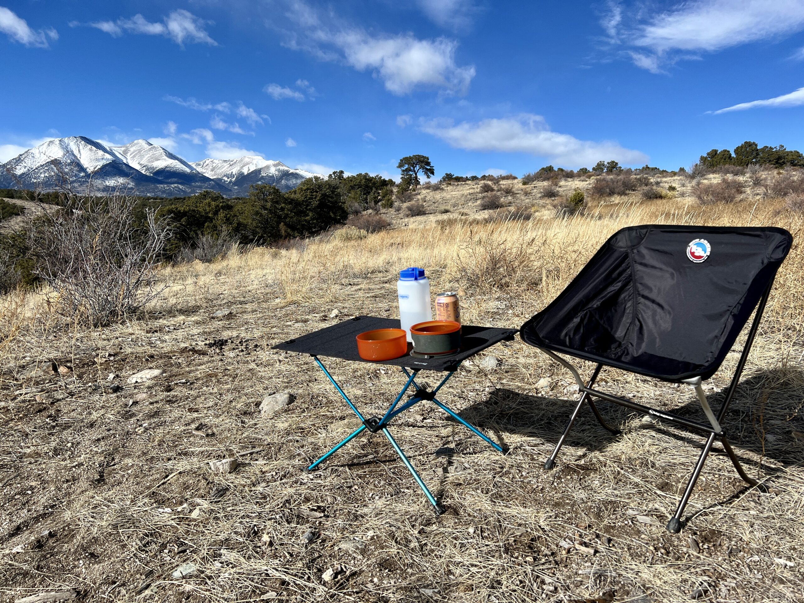 The Helinox One Hard Top Table is standing on dirt and scrub next to an empty black chair. It has orange cookware on it and a Nalgene bottle. The background is a scenic mountainscape.